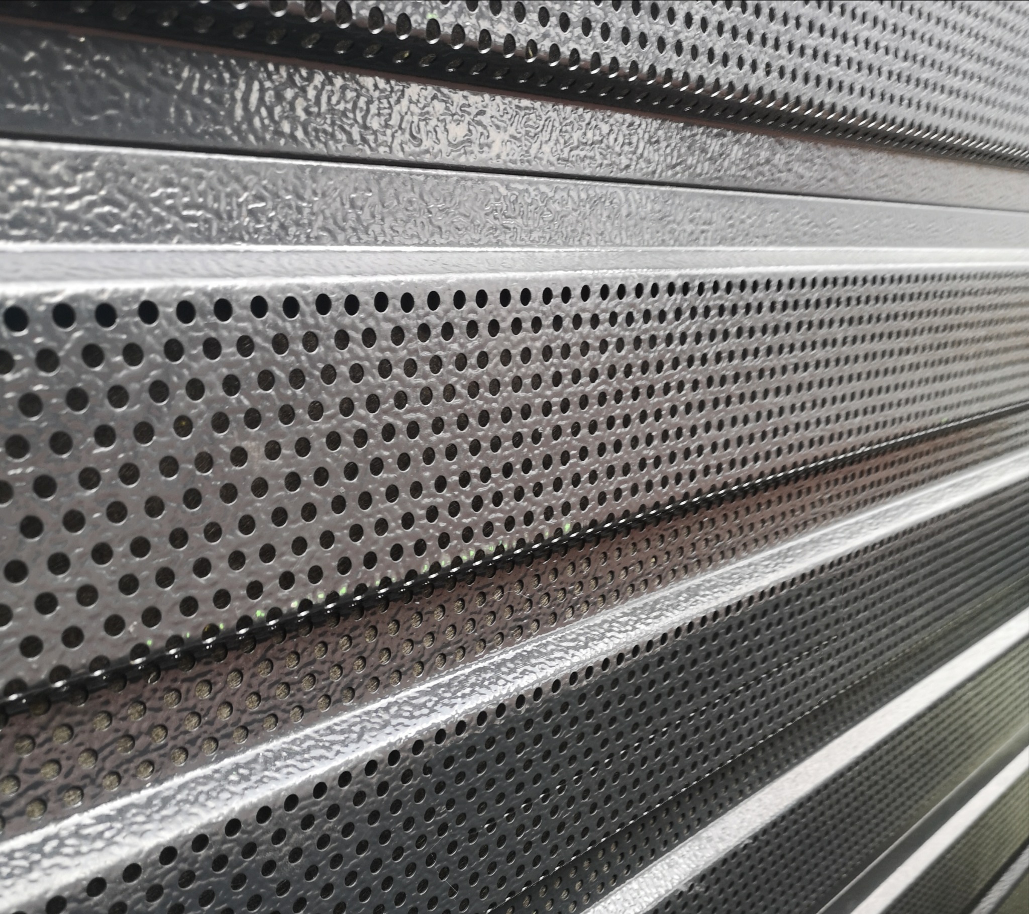 Szare, perforowane, stalowe profile ułożone warstwowo pod kątem, z widoczną fakturą i odbiciami światła.