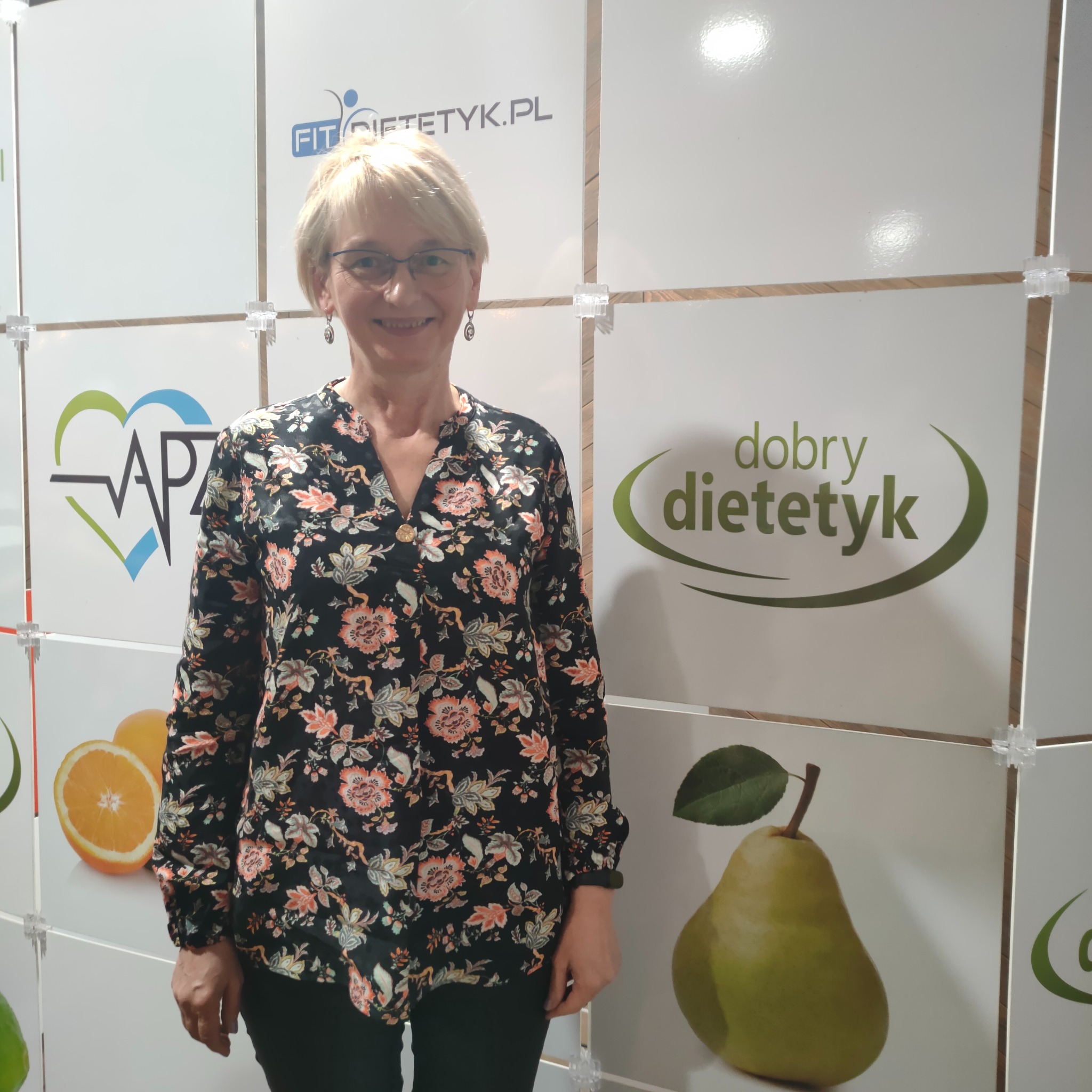 Uśmiechnięta dietetyczka w okularach, stojąca na tle ściany z logo firm dietetycznych i grafikami owoców.