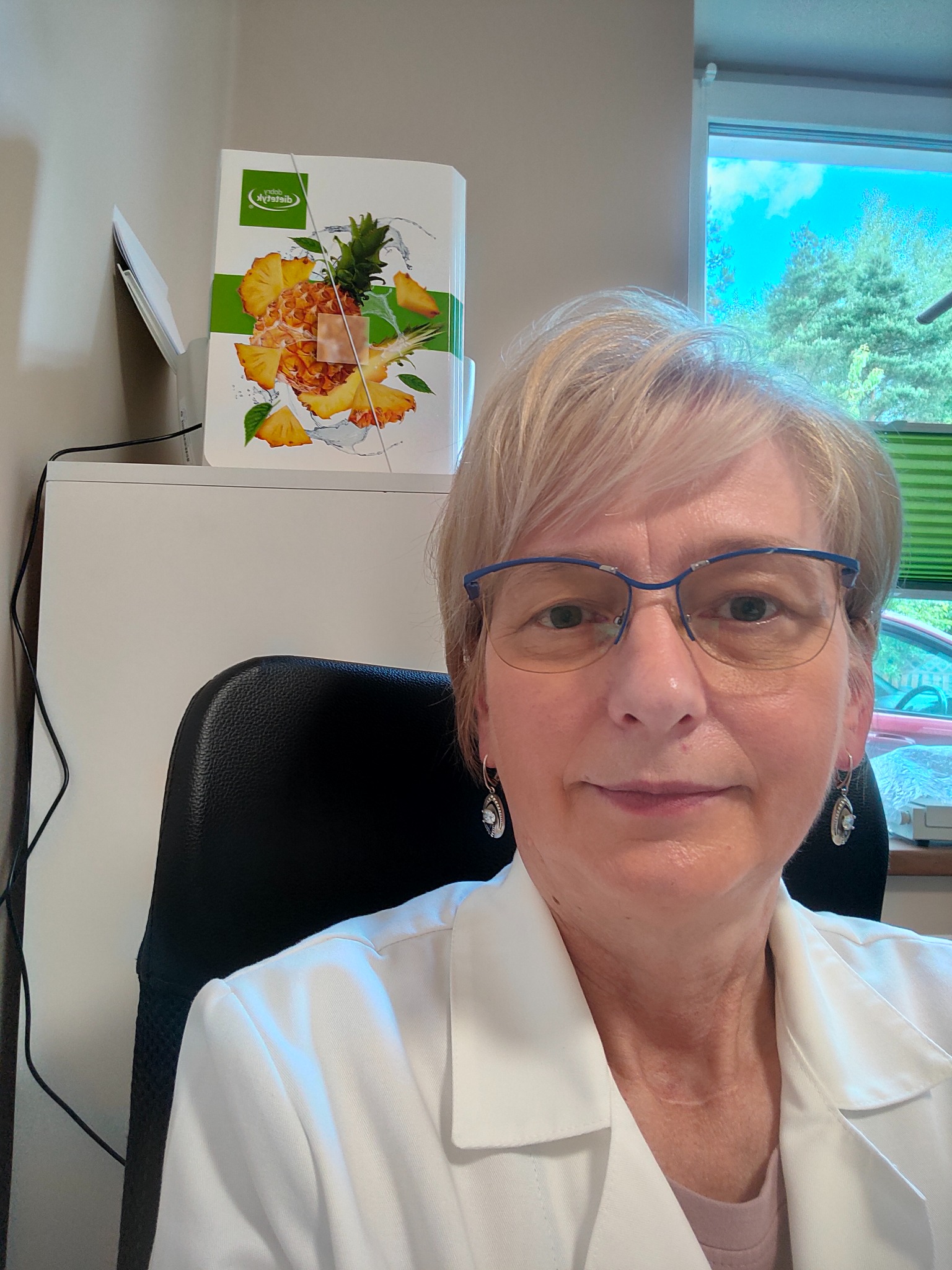 Portret kobiety w okularach i białym fartuchu lekarskim, siedzącej na czarnym krześle; w tle biała szafka z pudełkiem z motywem ananasa oraz okno z zielonymi żaluzjami.