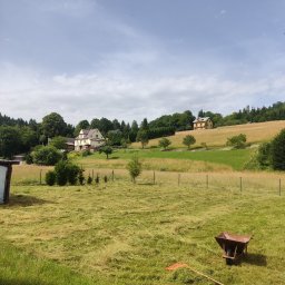 USŁUGI OGRODNICZE - Sielski krajobraz: świeżo skoszona trawa na polu, z widoczną grabiami i taczką, na wzgórzu domy otoczone zielenią, błękitne niebo z chmurami.