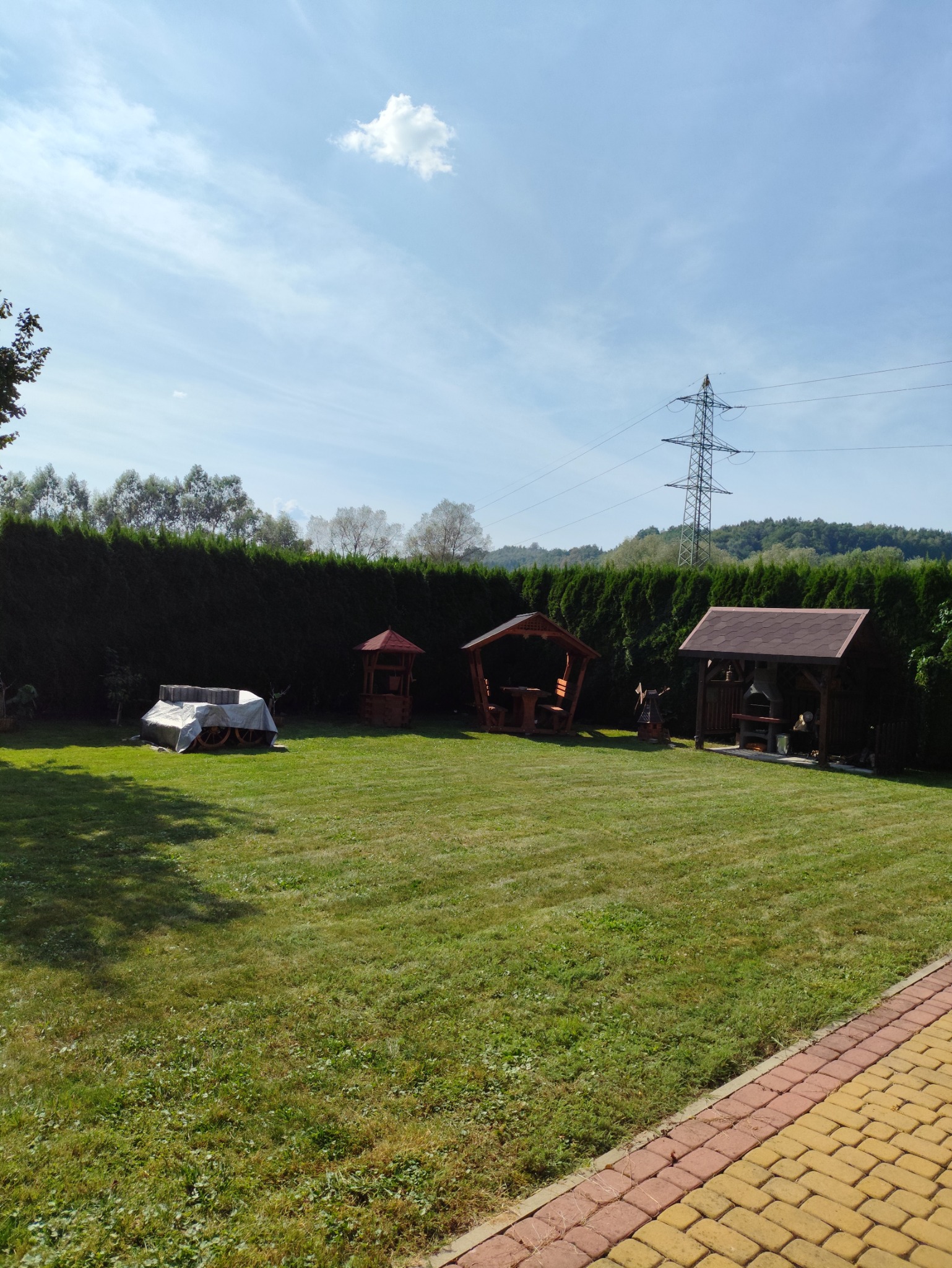 Zadbany trawnik z trzema drewnianymi altanami o różnych rozmiarach, wysoki żywopłot w tle i słup wysokiego napięcia na horyzoncie.