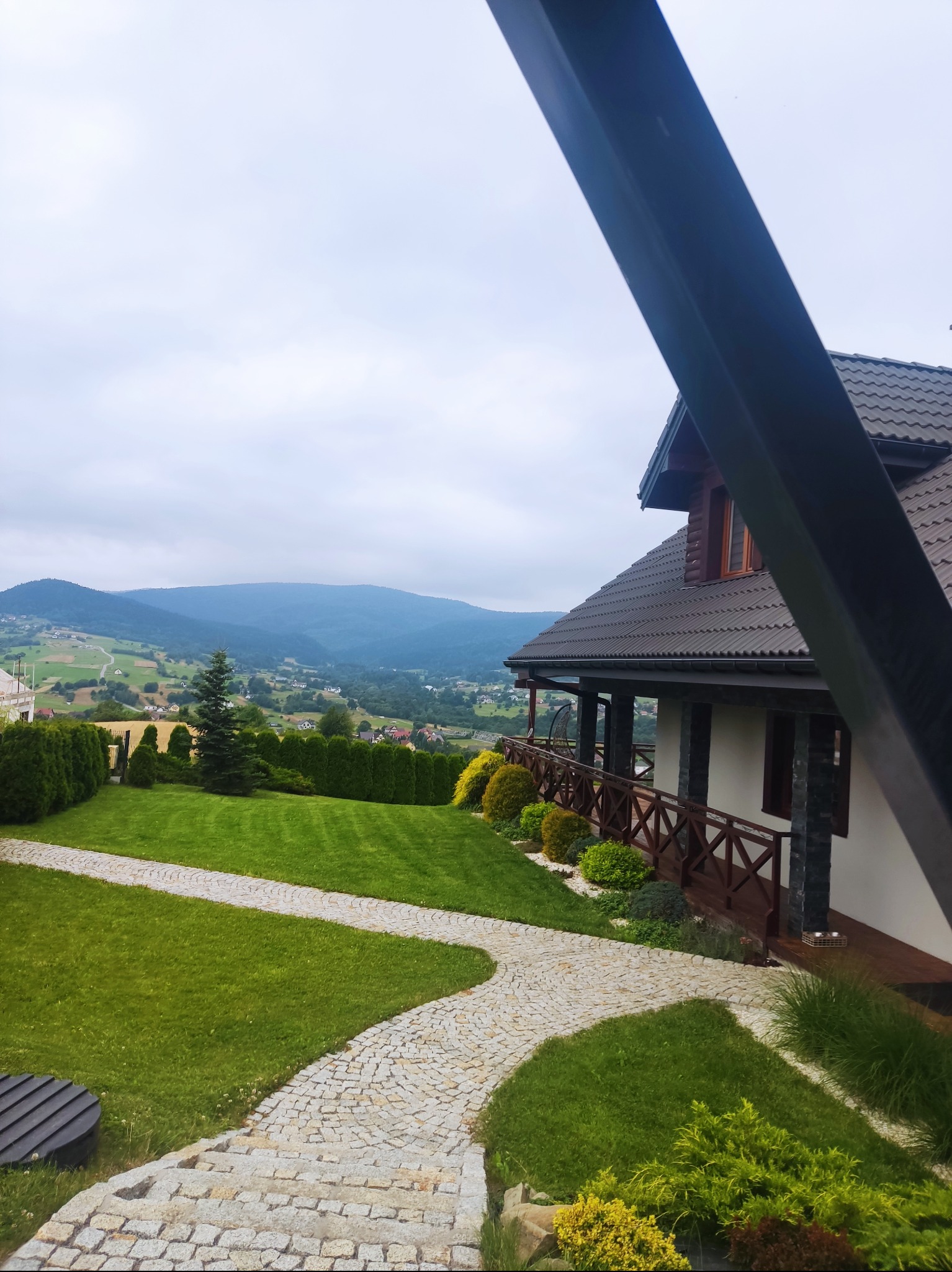 Ujęcie z perspektywy ogrodu z krętą ścieżką z kostki prowadzącą do domu z drewnianą balustradą, w tle rozległy widok na zalesione wzgórza pod pochmurnym niebem.