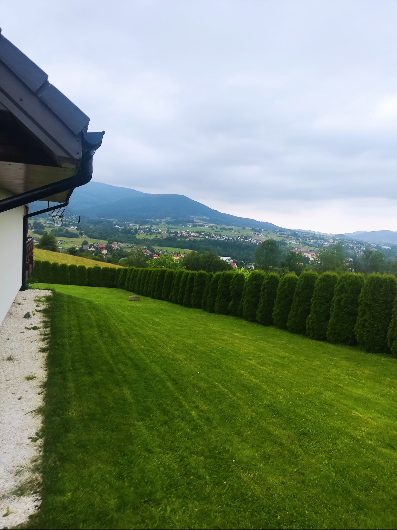 Ujęcie z boku domu na równo przystrzyżony trawnik, z równą linią żywopłotu z tuj, w tle zabudowa wiejska i góry pod pochmurnym niebem.