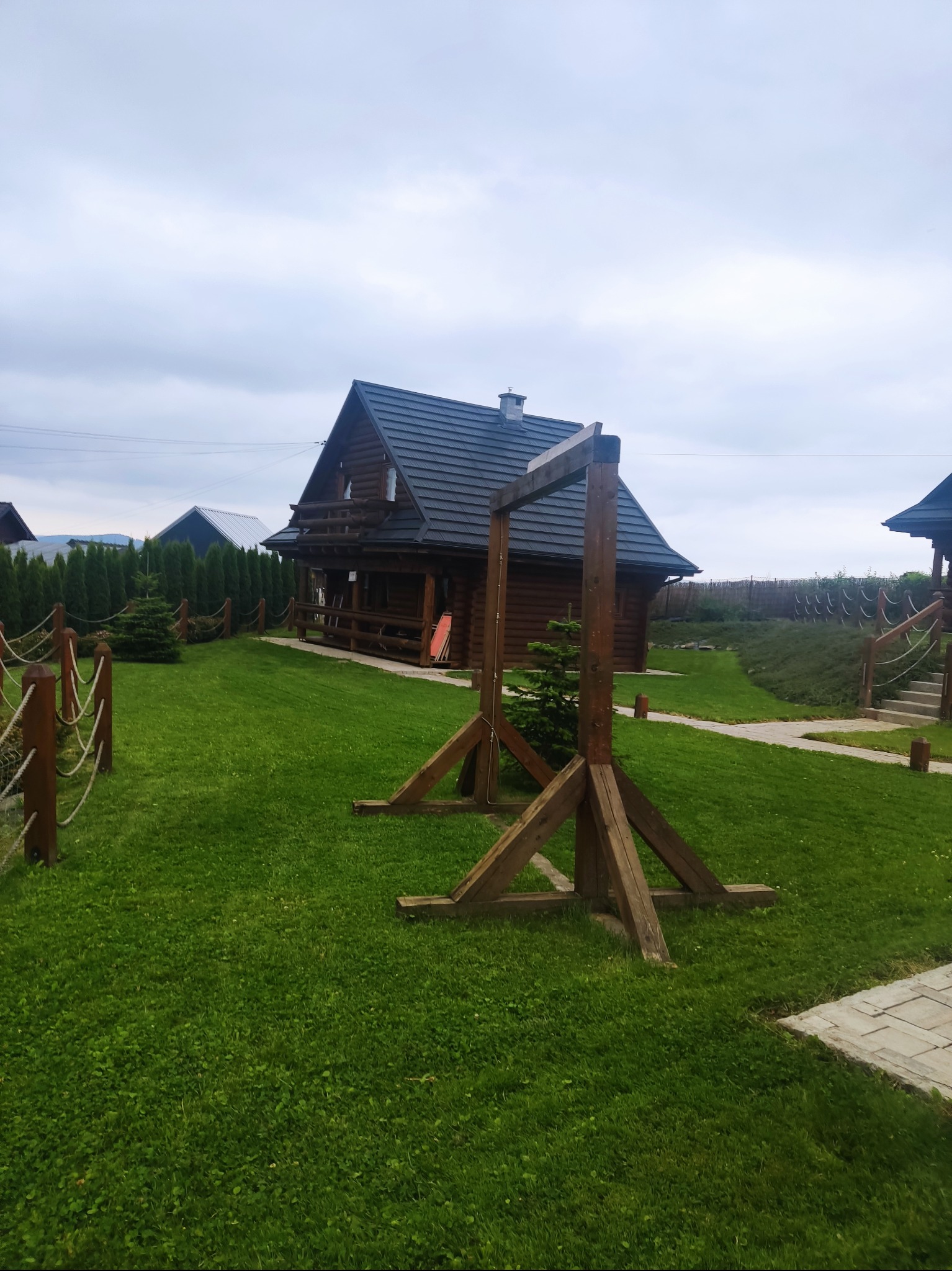 Rozległy trawnik przed drewnianym domem z bali, z drewnianą konstrukcją w kształcie bramy na pierwszym planie i ozdobnym ogrodzeniem z lin.