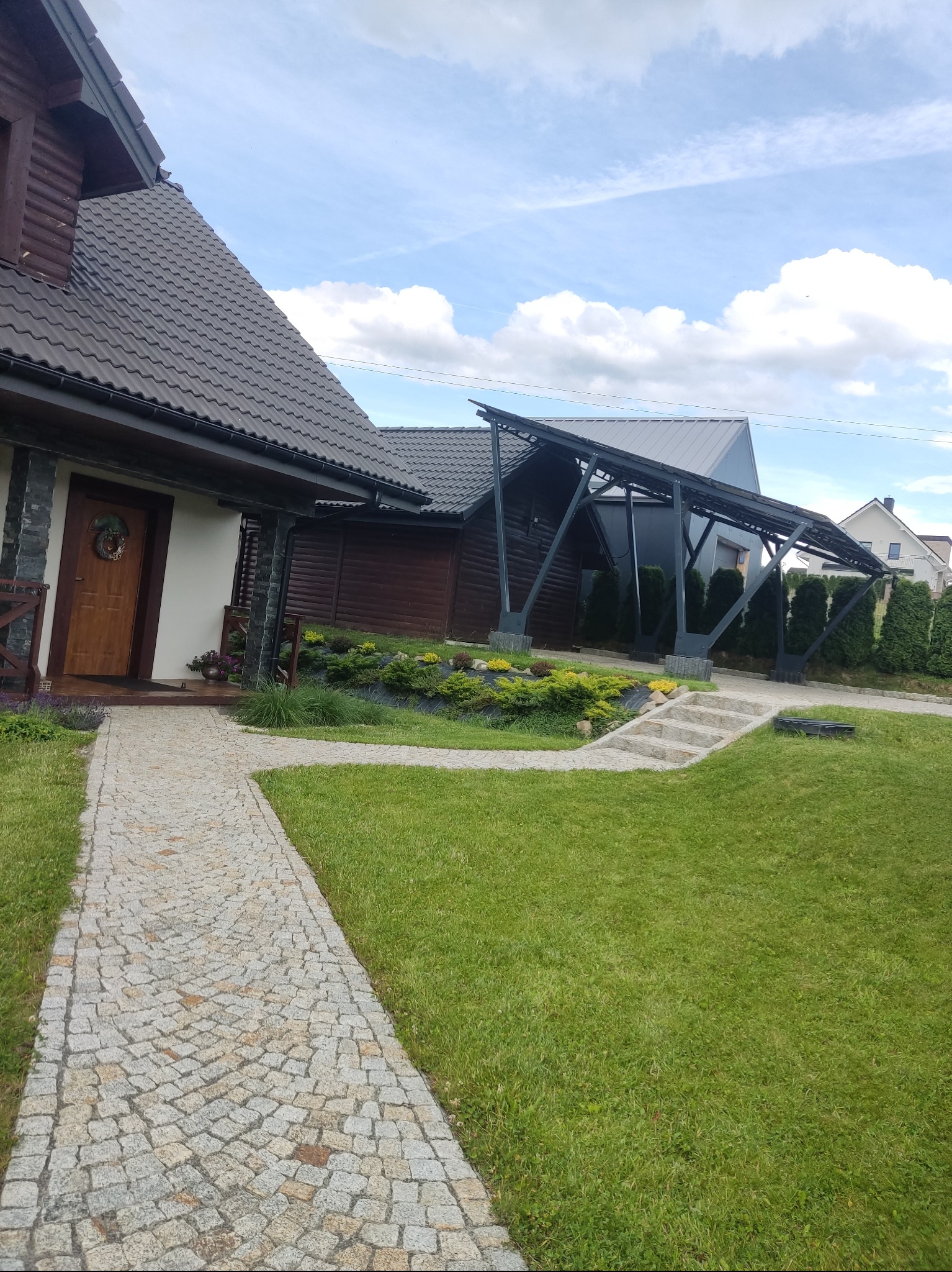 Ujęcie z poziomu trawnika na zadbany ogród przydomowy z kamiennym chodnikiem, rabatami kwiatowymi i strzyżonymi krzewami, na drugim planie dom z drewnianą elewacją i zadaszone miejsce parkingowe.