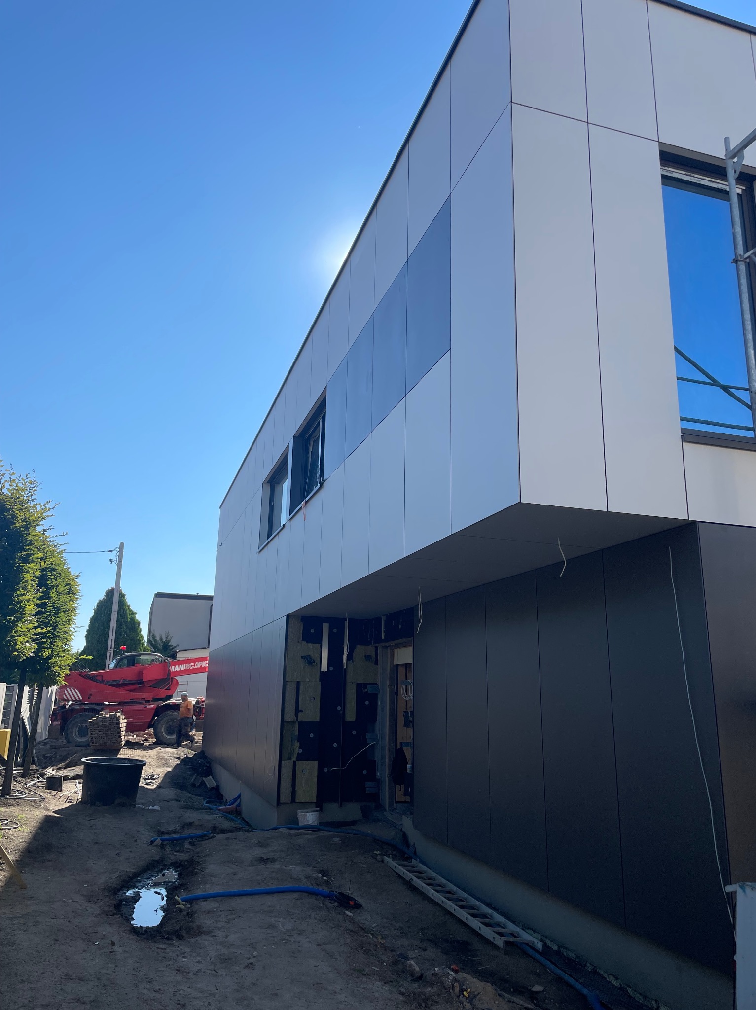 Nowoczesny budynek w trakcie prac elewacyjnych, widoczne panele w kolorze białym i grafitowym, rusztowania, podnośnik teleskopowy Maniscopic w tle, teren budowy z kałużami.