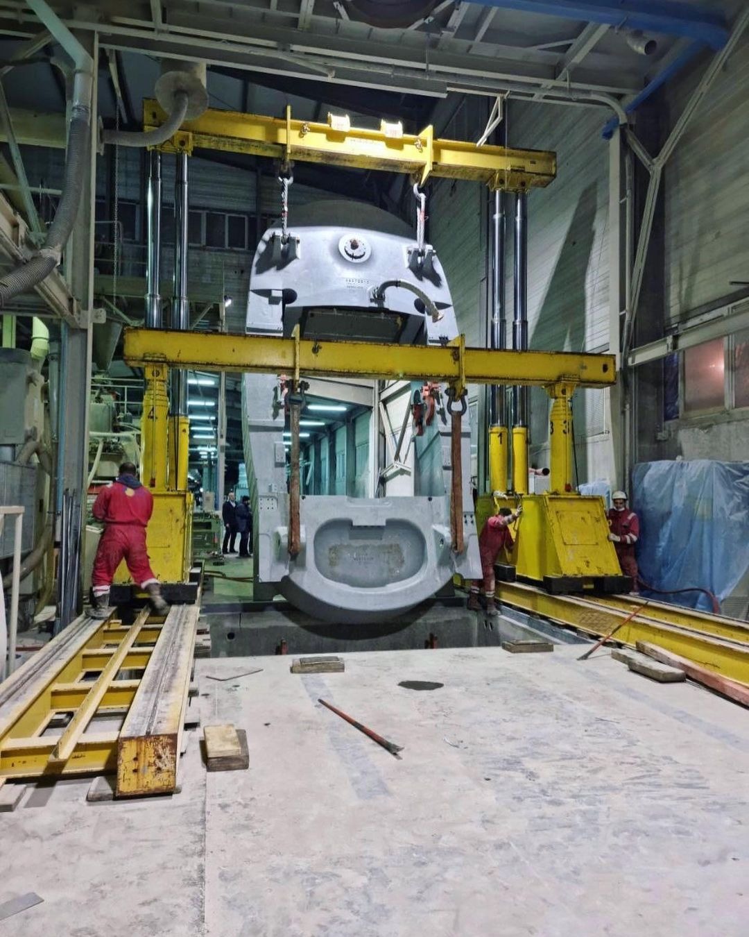 Montaż formy odlewniczej za pomocą suwnicy w hali przemysłowej, pracownicy w kombinezonach kontrolują proces.