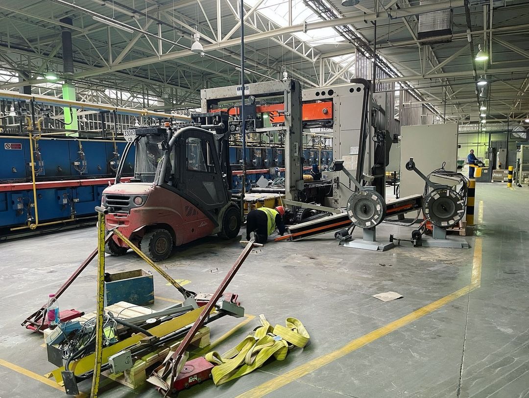 Wnętrze hali przemysłowej z wózkiem widłowym Linde i pracownikiem w żółtej kamizelce obsługującym maszynę do obróbki metalu; na podłodze narzędzia i akcesoria.