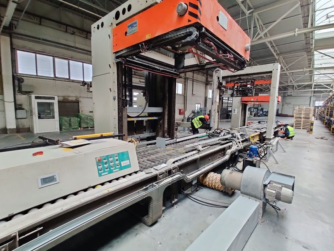 Wnętrze hali produkcyjnej z suwnicą bramową i linią transportową; pracownicy w kamizelkach odblaskowych kontrolują system