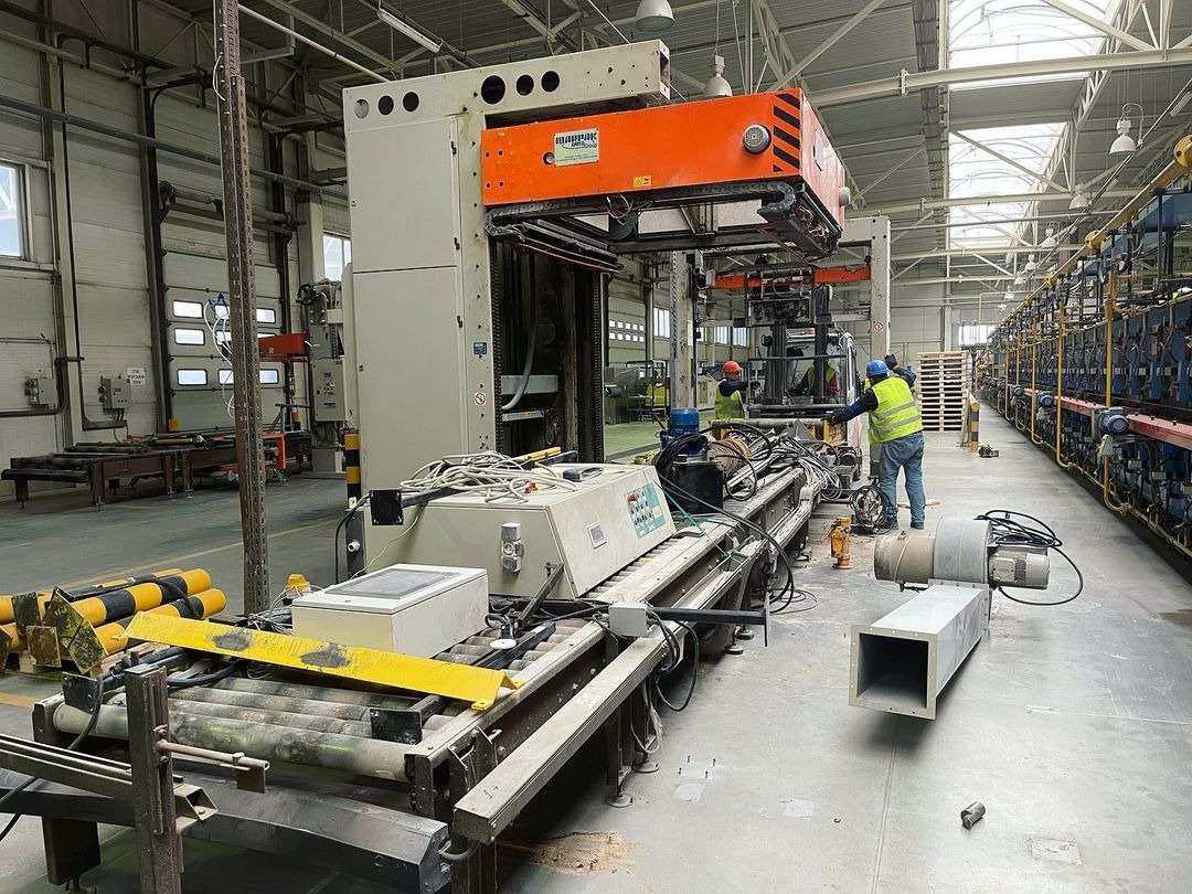 Wnętrze hali produkcyjnej z suwnicą bramową i pracownikami w kamizelkach odblaskowych, transportującą palety z gotowymi produktami, widoczne regały z produktami w tle