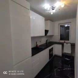 Nowoczesna kuchnia z białymi, błyszczącymi frontami szafek, czarnym blatem i podłogą z czarnego marmuru z białymi żyłami; okno z rośliną i krzesłem w tle.