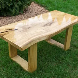 Stolik kawowy z blatem z litego drewna o nieregularnych krawędziach, lakierowany na wysoki połysk, na tle zielonego trawnika.