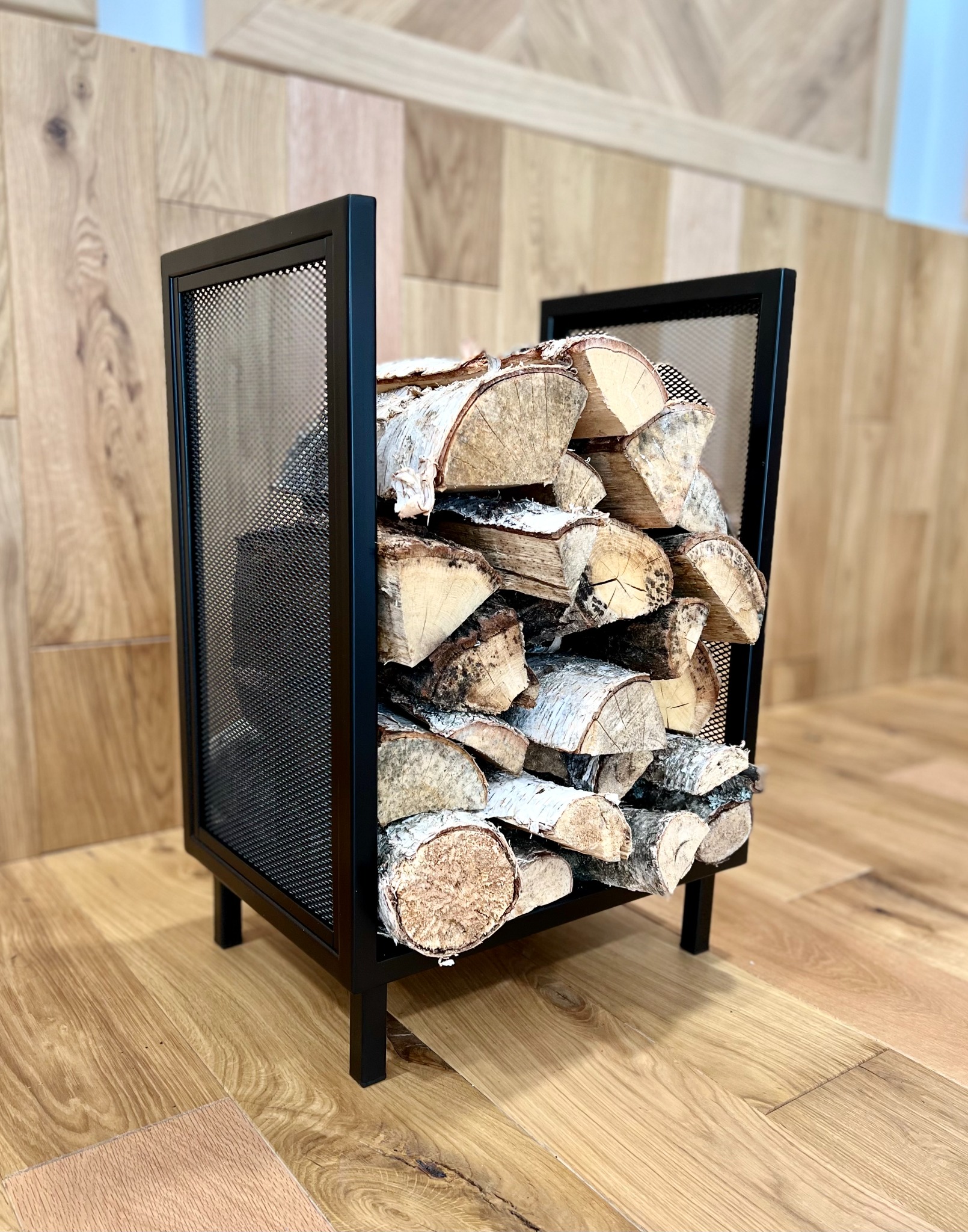 Metalowy stojak na drewno kominkowe, wypełniony pociętymi kawałkami drewna brzozowego, na drewnianej podłodze.