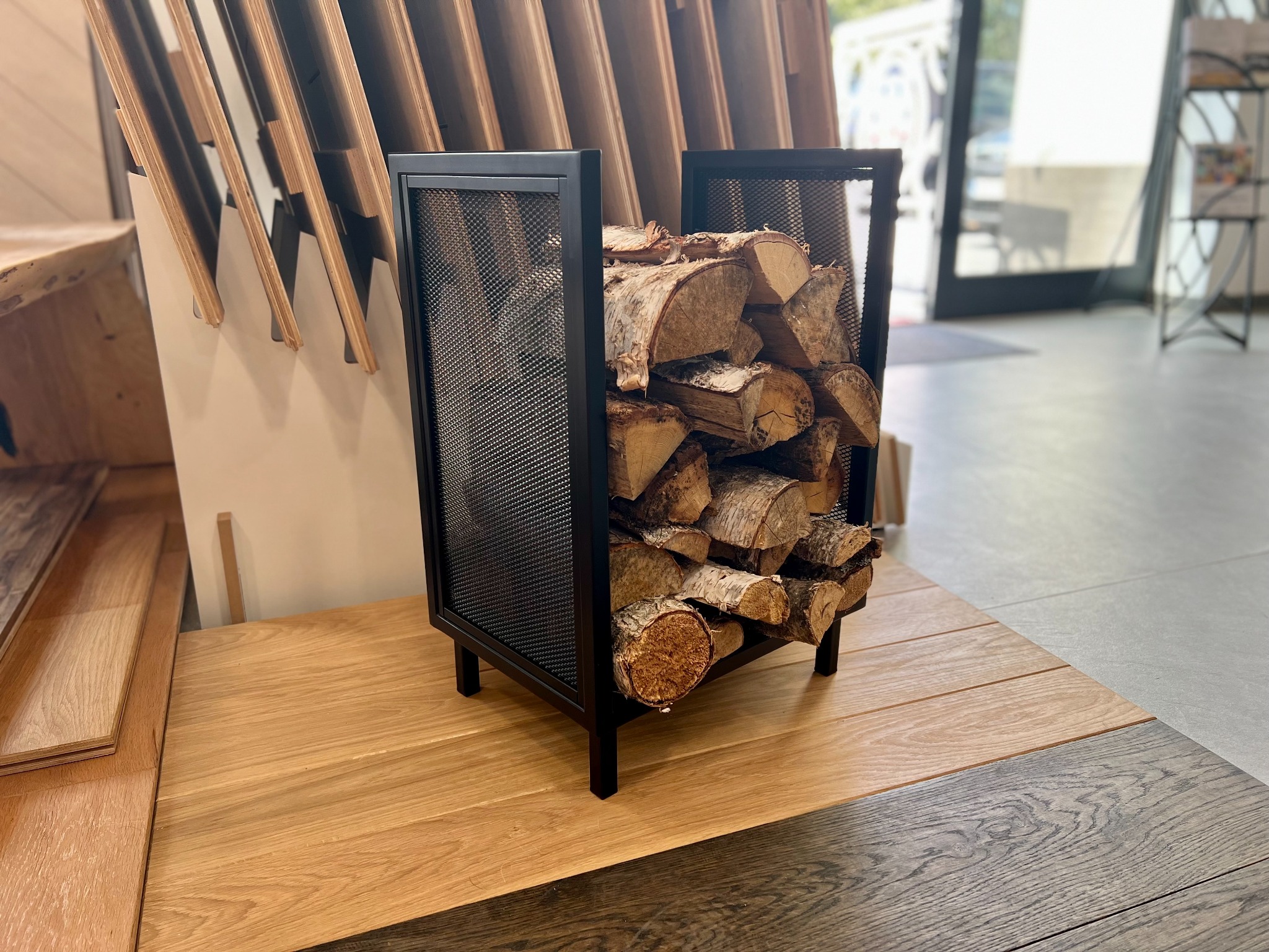 Metalowy kosz z siatki wypełniony drewnem opałowym stoi na drewnianym blacie w salonie ekspozycyjnym, w tle widoczne próbki podłóg i drzwi.