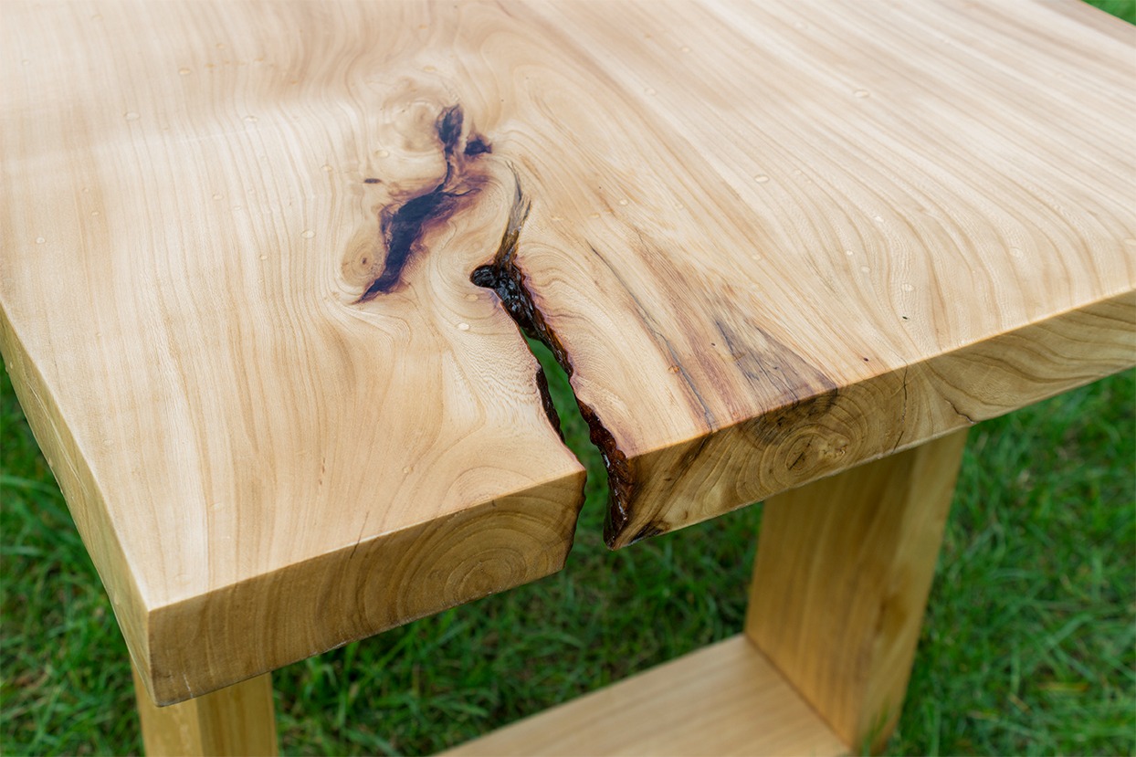 Blat drewniany z widocznym usłojeniem i naturalną szczeliną wypełnioną ciemną żywicą, osadzony na drewnianych nogach, na tle zielonej trawy.