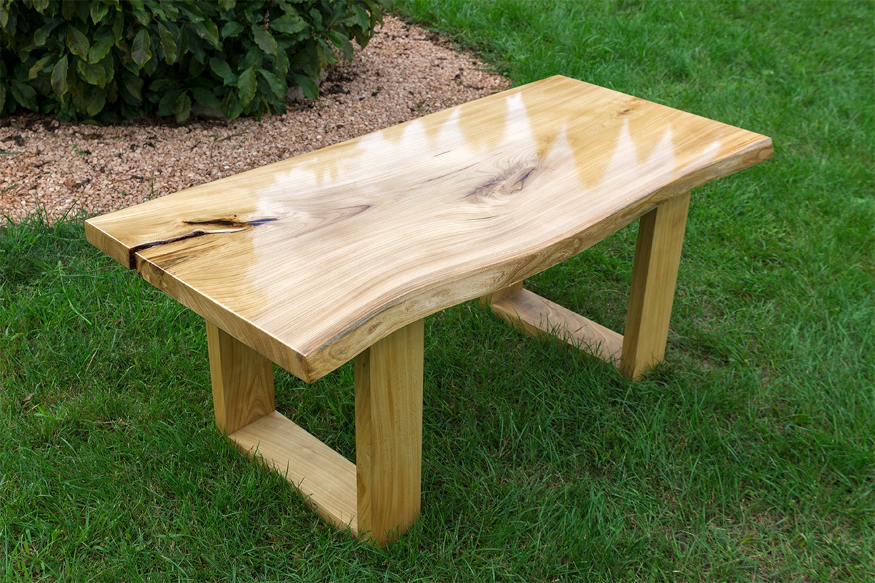 Stolik kawowy z blatem z litego drewna o nieregularnych krawędziach, lakierowany na wysoki połysk, na tle zielonego trawnika.