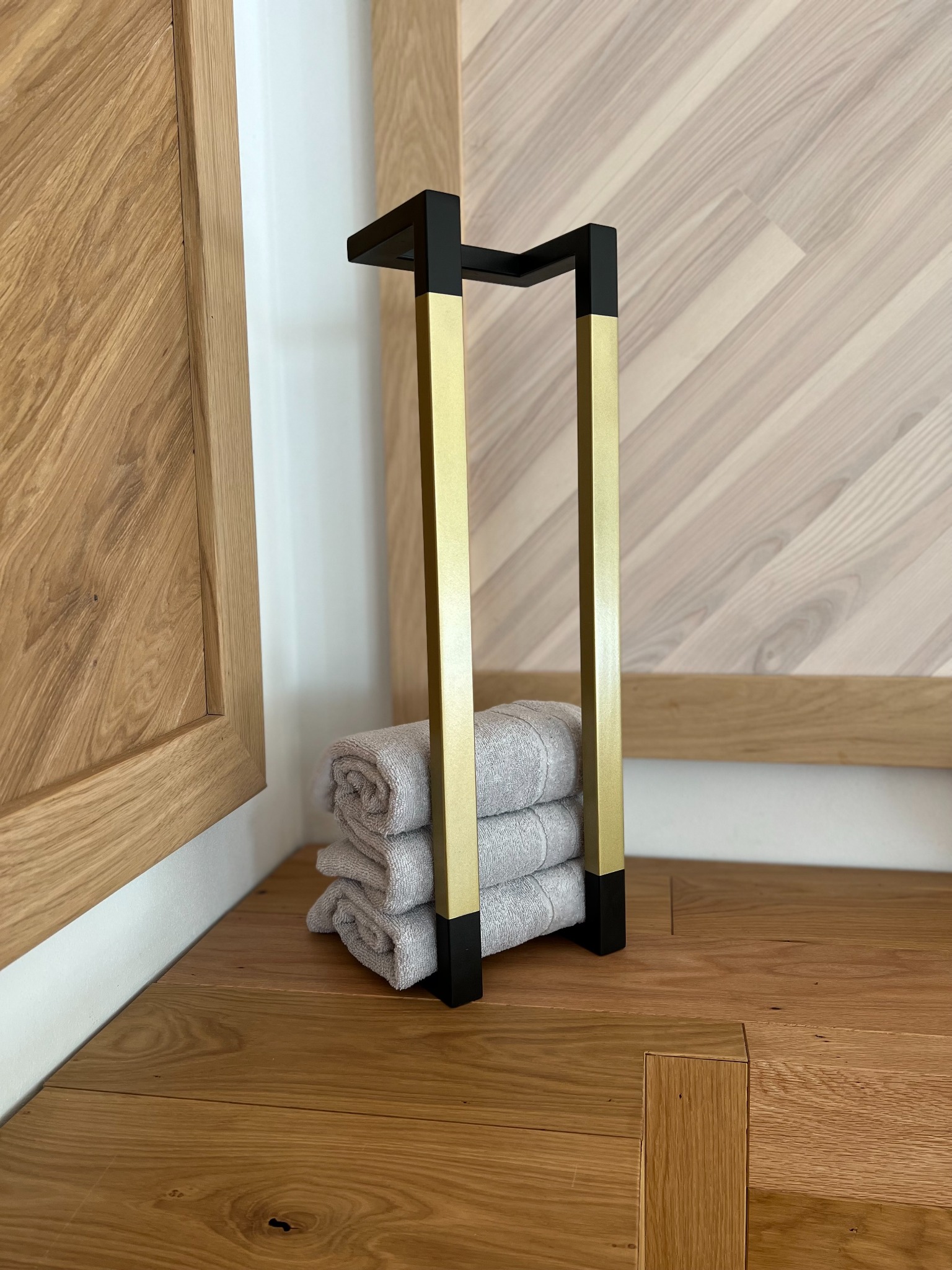 Stojak na ręczniki z metalu (złoto i czerń) z trzema złożonymi ręcznikami na podłodze z drewnianych paneli, widoczne fragmenty drewnianej zabudowy ściany.