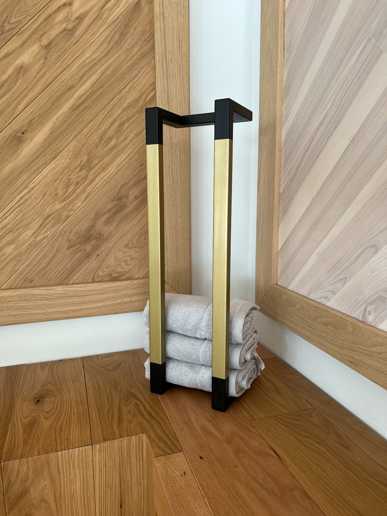 Stojak na ręczniki z metalu w kolorach złota i czerni, ustawiony na drewnianej podłodze, z trzema zwiniętymi ręcznikami w środku, na tle drewnianych paneli ściennych.