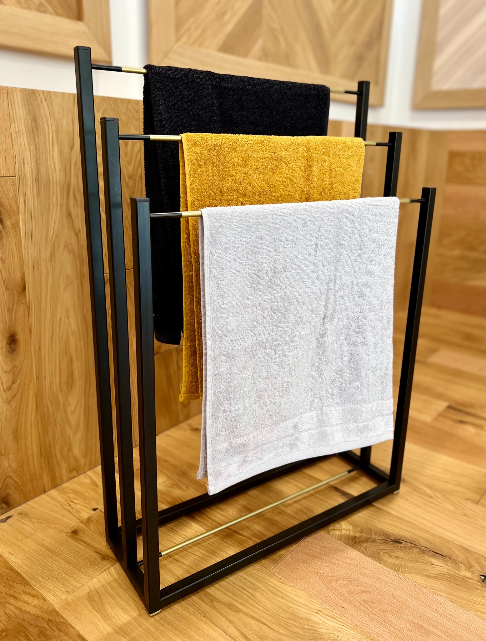 Wolnostojący, nowoczesny wieszak na ręczniki w kolorach czerni i złota, z trzema ręcznikami: czarnym, musztardowym i białym na tle drewnianej podłogi i ściany.