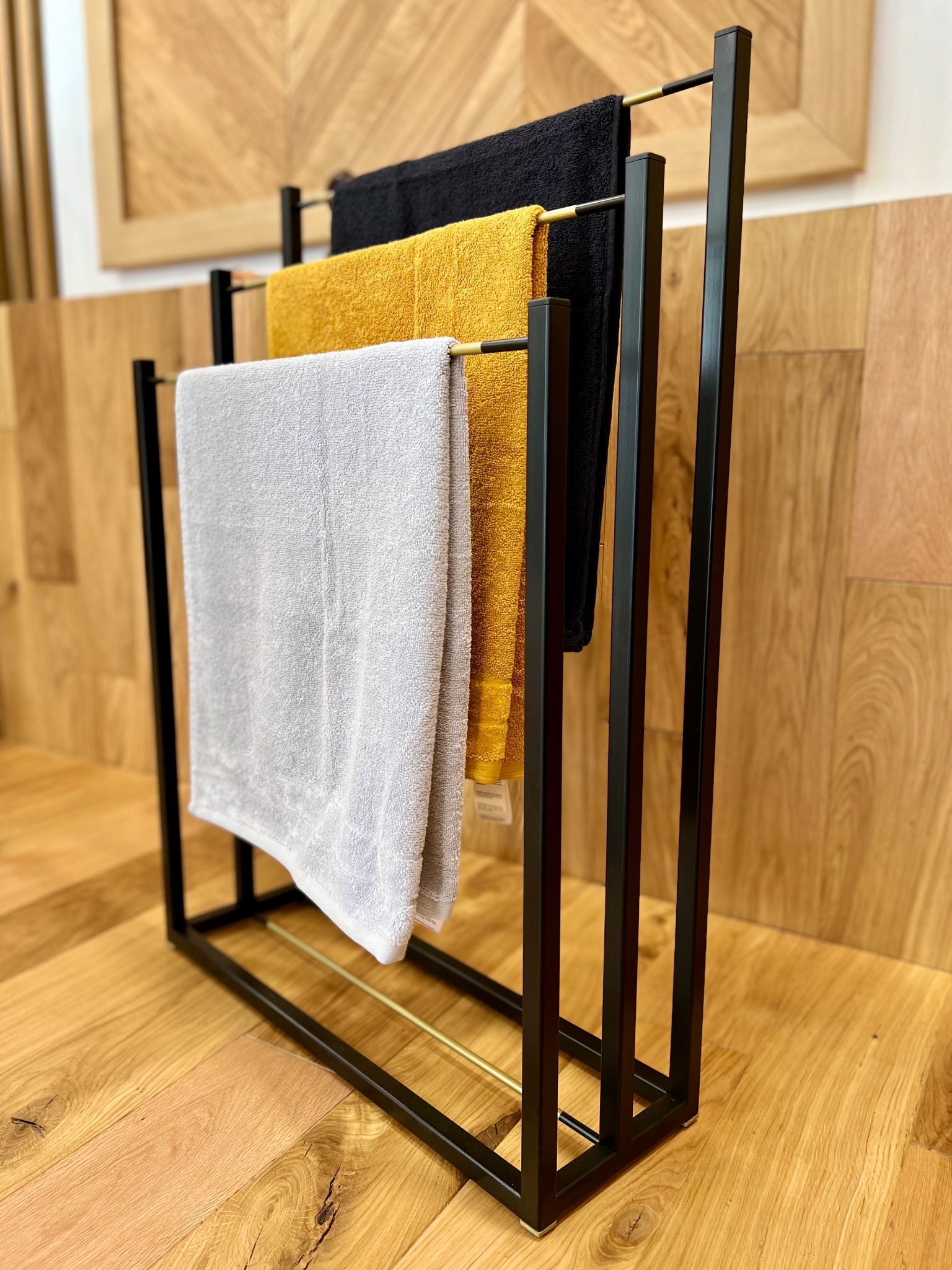 Metalowy stojak na ręczniki w stylu loft z trzema ręcznikami: szarym, żółtym i czarnym, na tle drewnianej ściany i podłogi z parkietu.
