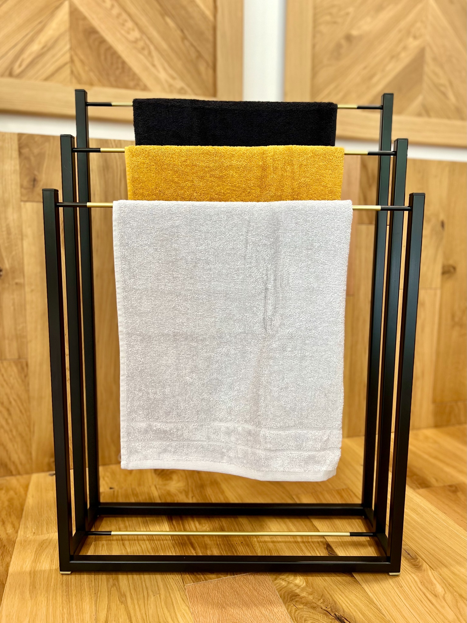 Czarny, złoty i biały ręcznik zawieszone na czarnym, metalowym stojaku z elementami w kolorze złota na tle drewnianej ściany i podłogi.