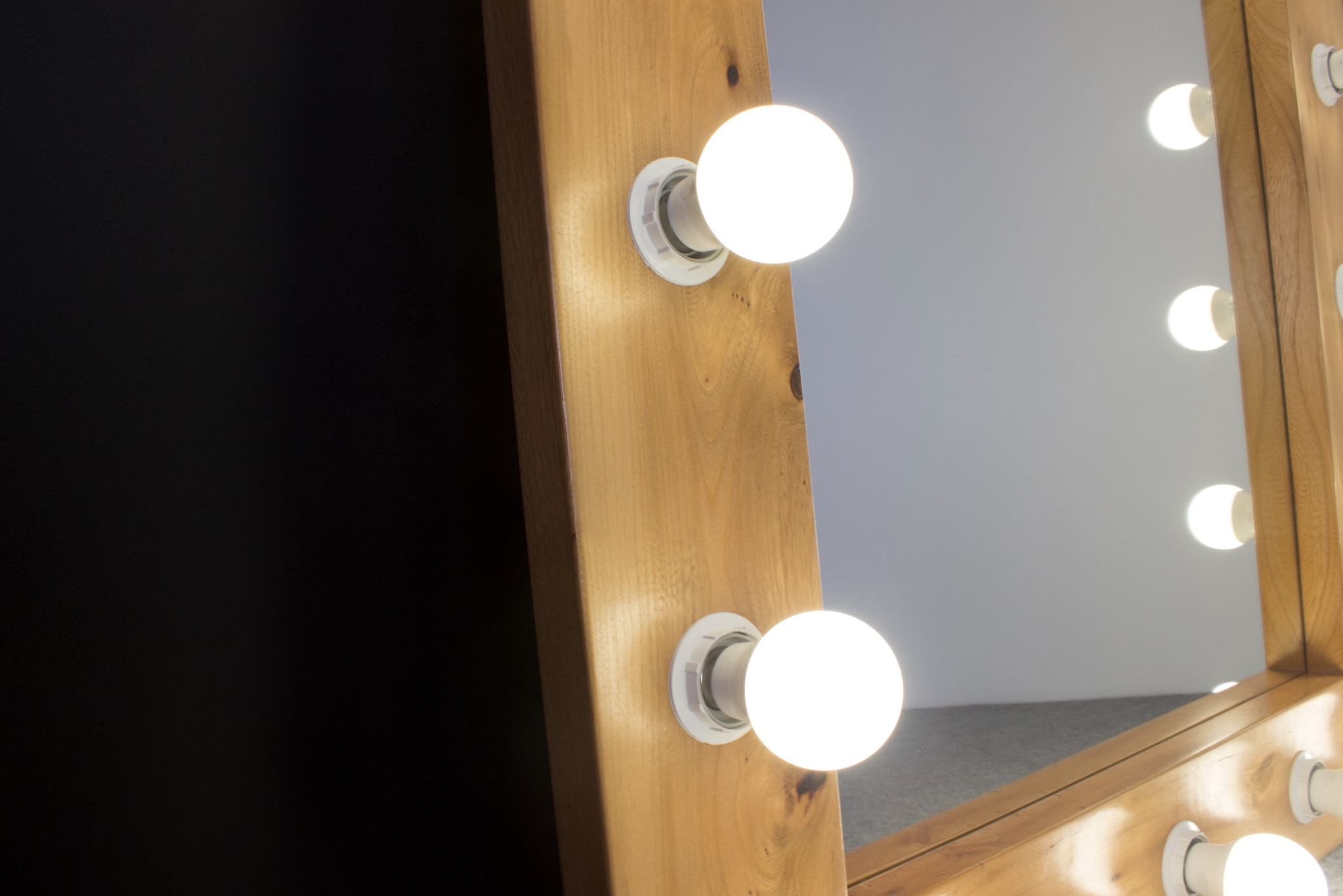 Fragment lustra do makijażu z drewnianą ramą i okrągłymi żarówkami, widoczne odbicie w lustrze i czarne tło po lewej stronie.