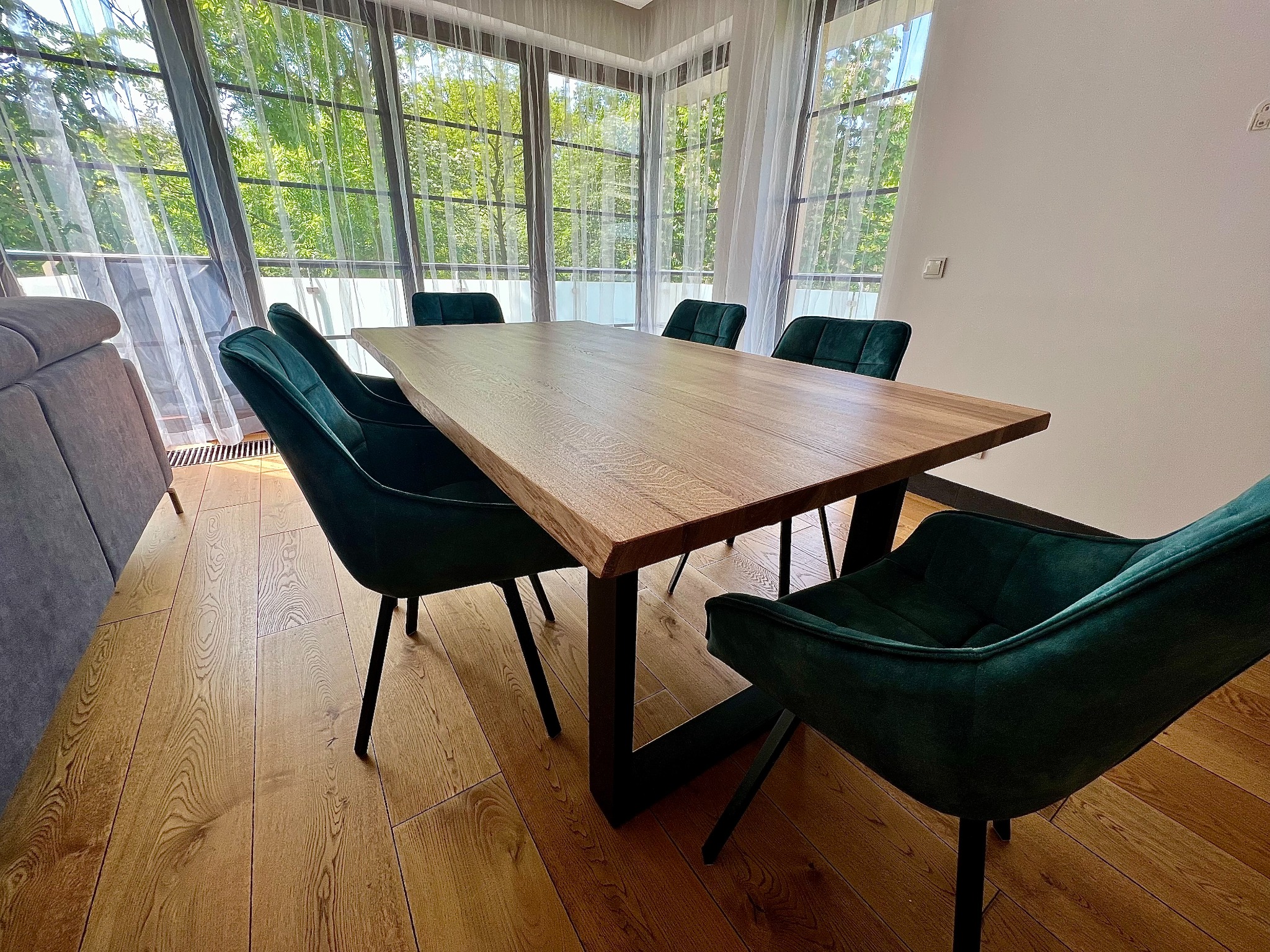 Jadalnia z drewnianym stołem o surowym wykończeniu i krzesłami tapicerowanymi ciemnozielonym welurem, widok na zieleń za oknem.