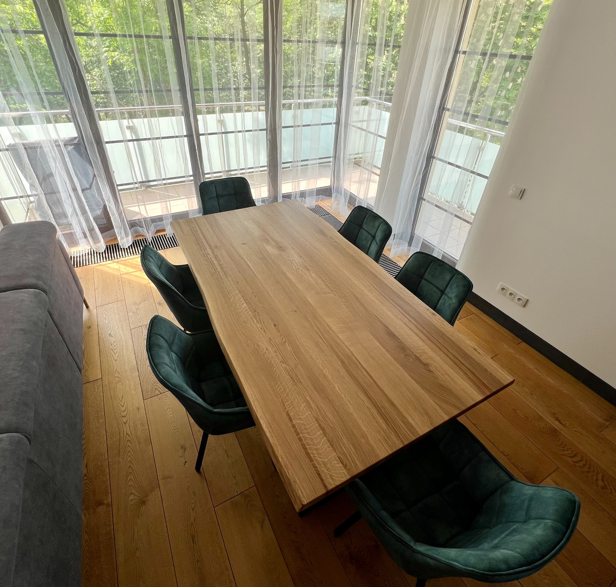 Drewniany stół z blatem o nieregularnych krawędziach w nowoczesnym wnętrzu z zielonymi krzesłami i widokiem na balkon przez zasłonięte okno.