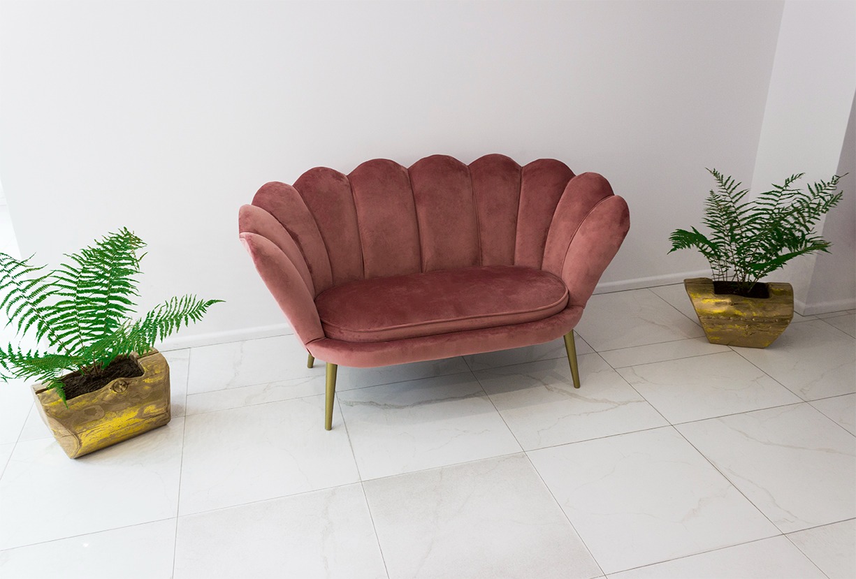 Różowa, welurowa sofa w kształcie muszelki na złotych nóżkach, ustawiona na białych płytkach, z paprotkami w złotych doniczkach po bokach.
