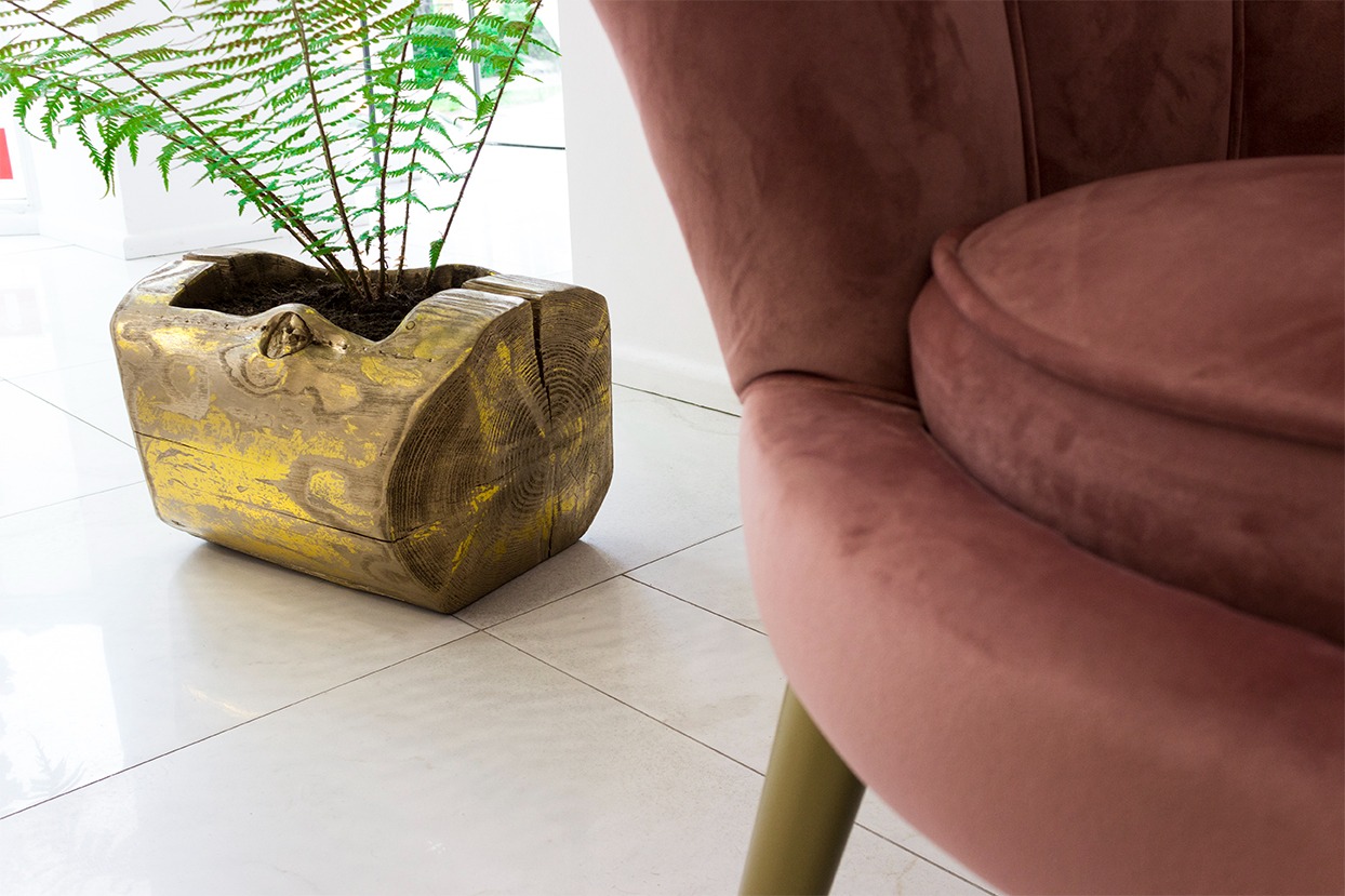 Dekoracyjny pień drzewa z paprotką jako doniczka, obok fragmentu różowego, welurowego fotela na tle białej podłogi.