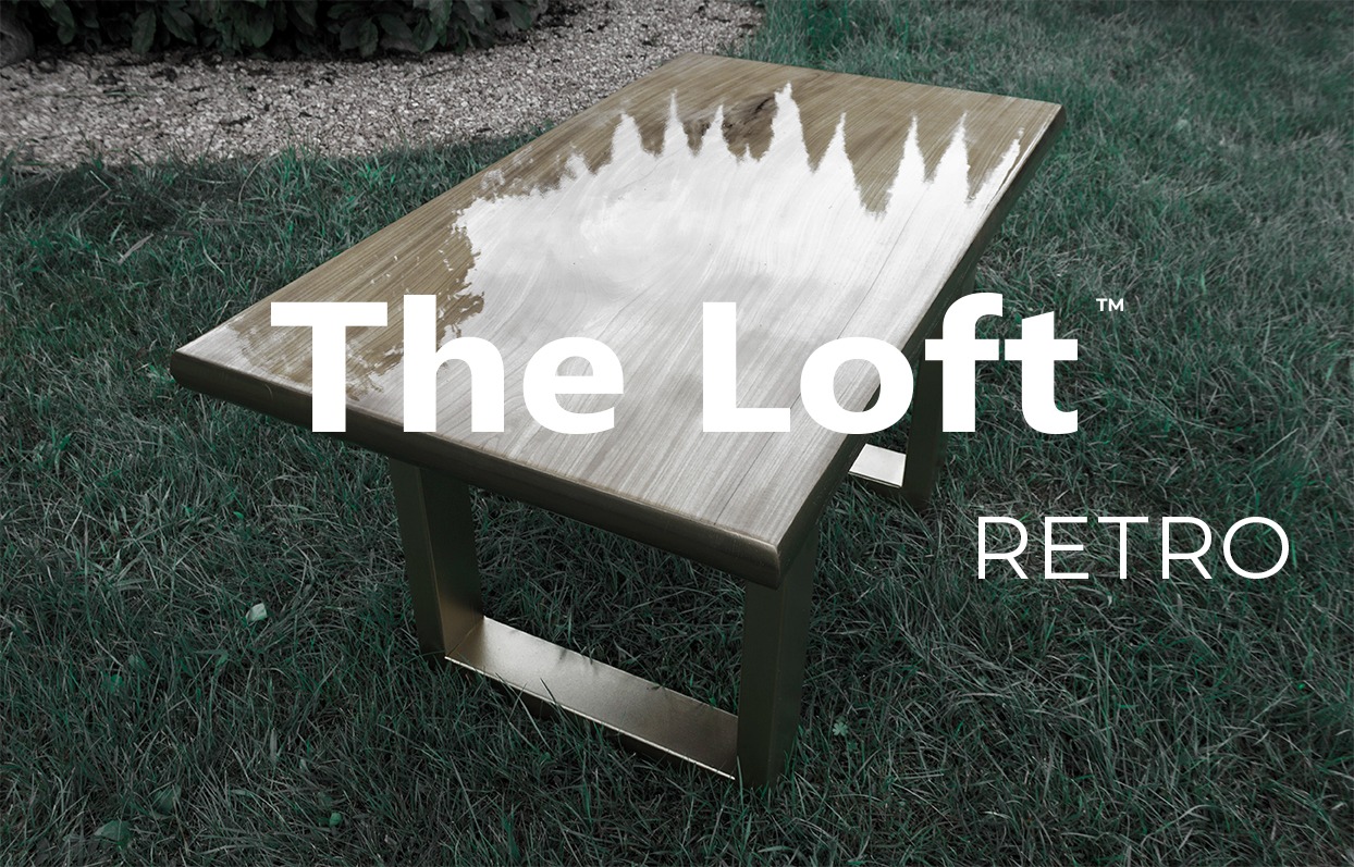 Stół z blatem z jasnego drewna, z metalowymi nogami w stylu loft retro, umieszczony na trawie, z widocznym odbiciem nieba w lakierowanej powierzchni blatu. Na zdjęciu widoczne napisy 'The Loft'...