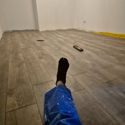 PL HANDYMAN TRANSPORT AND CONSTRUCTION PAWEŁ LEBODA - Ułożone panele podłogowe w szarym odcieniu drewna, widok z perspektywy osoby w roboczym ubraniu z plamami farby, poziomica i nożyk na podłodze.