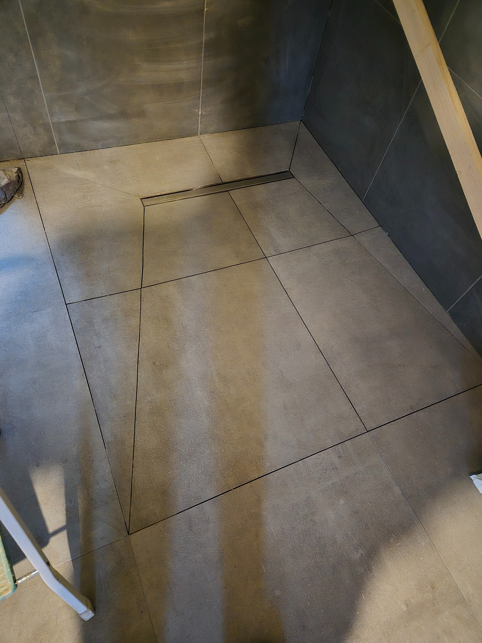 Wykończona podłoga w kabinie prysznicowej z odpływem liniowym, wykonana z szarych, matowych płytek o nieregularnym kształcie, ułożonych pod kątem.