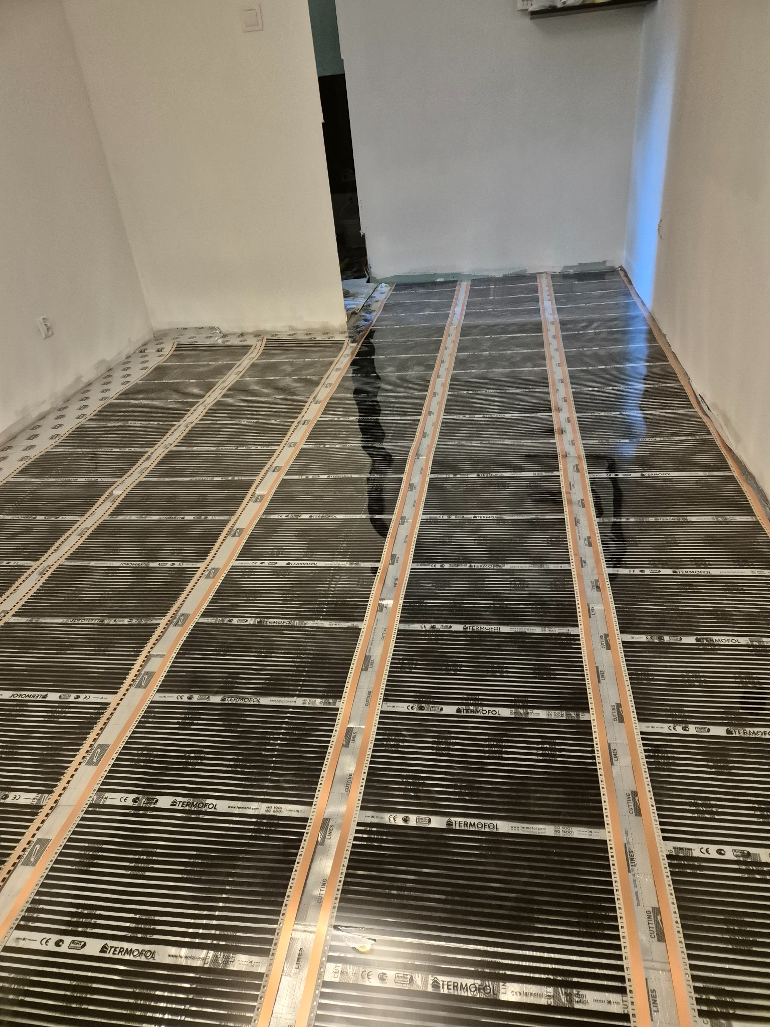 Montaż folii grzewczej Termofol na podłodze w jasnym pomieszczeniu. Widoczne równoległe pasy folii i taśmy montażowe. Ściany białe, surowe.