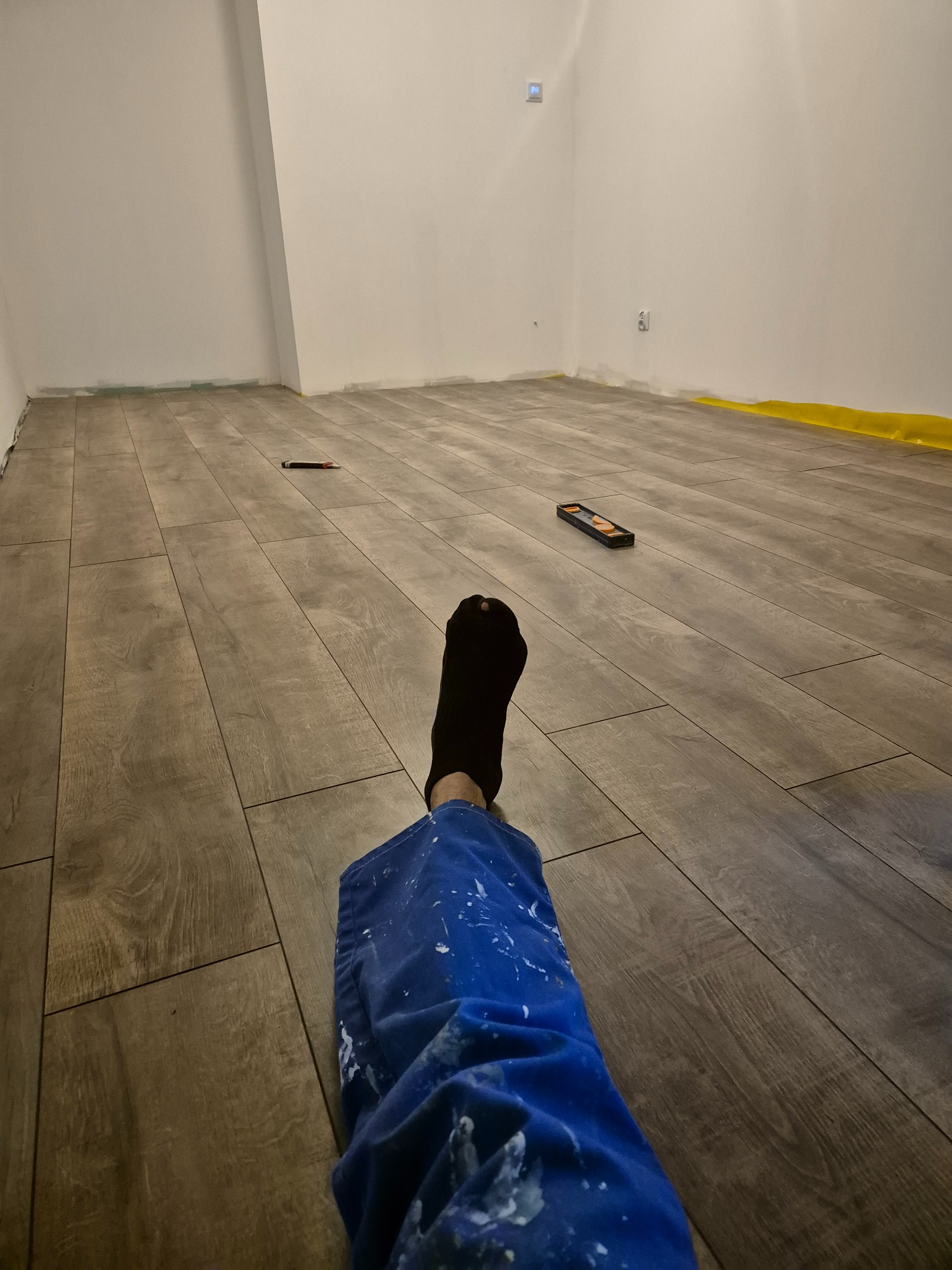 Ułożone panele podłogowe w szarym odcieniu drewna, widok z perspektywy osoby w roboczym ubraniu z plamami farby, poziomica i nożyk na podłodze.