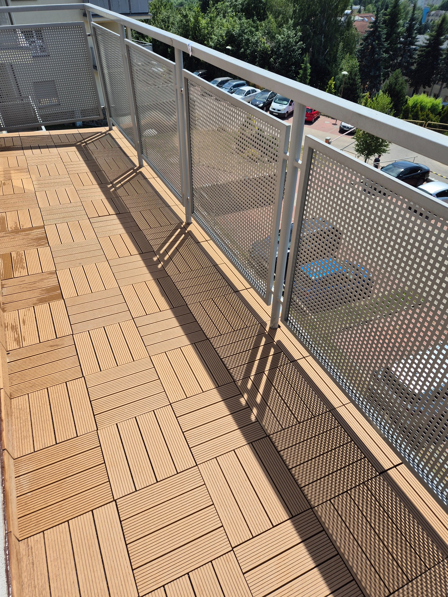 Balkon z nową, drewnopodobną podłogą z desek kompozytowych, wykończony metalową balustradą z perforowanymi panelami. Widok z góry, światło słoneczne.