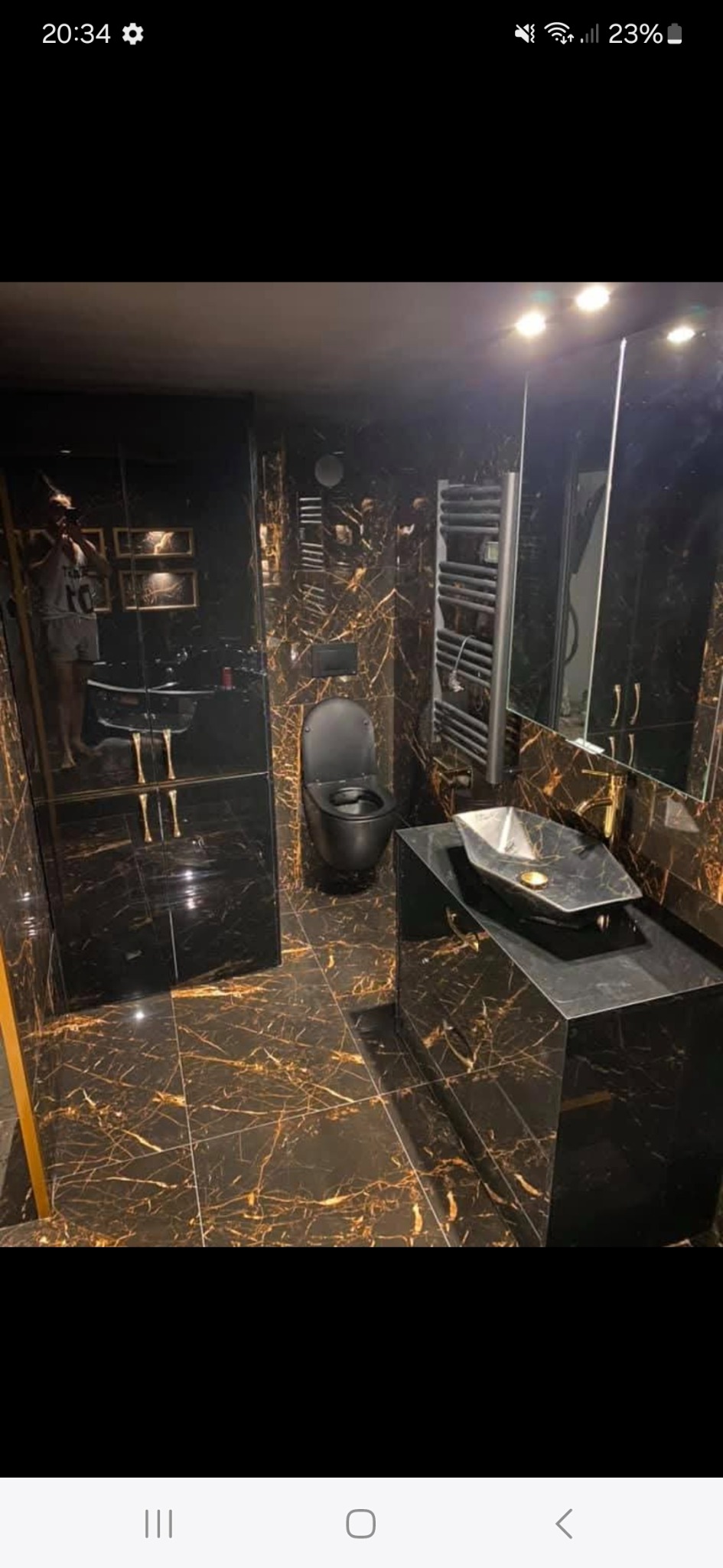 Elegancka łazienka z czarnymi płytkami imitującymi marmur ze złotymi żyłami, czarną armaturą i oświetleniem punktowym. W lustrze odbija się osoba robiąca zdjęcie.