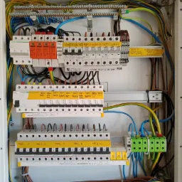 Otwarta skrzynka elektryczna z widocznymi zabezpieczeniami, przewodami i żółtymi etykietami opisującymi obwody, w tym 'Kontrola faz', 'Lampy', 'Pralnia' i 'Magiel'.