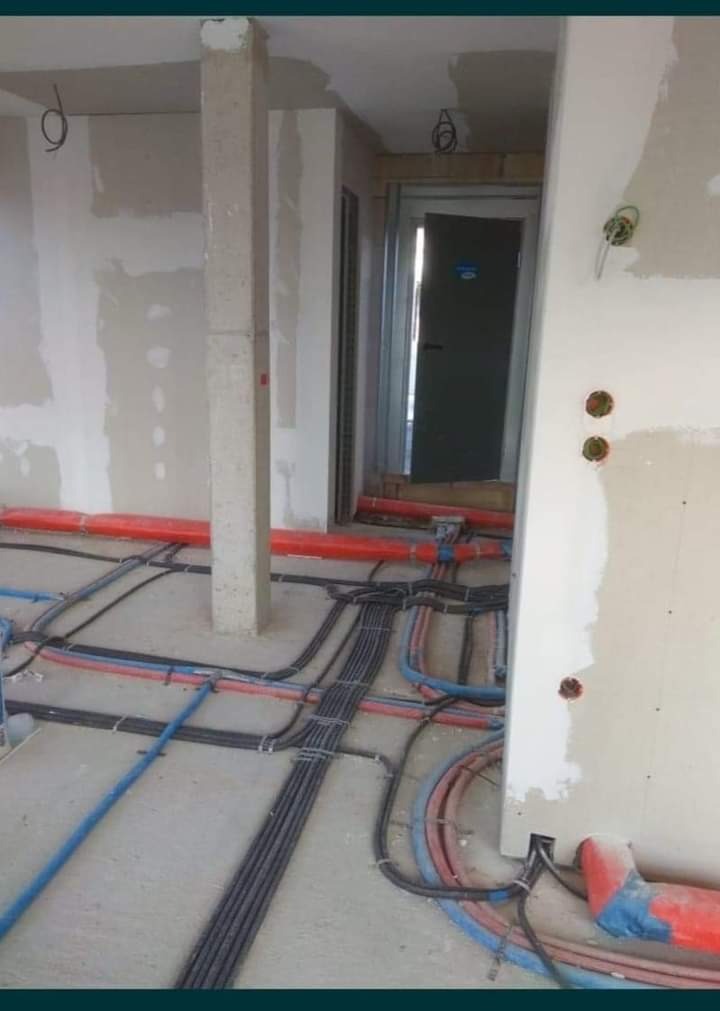 Instalacja elektryczna w surowym stanie: widoczne liczne przewody elektryczne i rury w kolorach czarnym, niebieskim i czerwonym ułożone na podłodze, ściany w trakcie wykończenia, surowy betonowy...