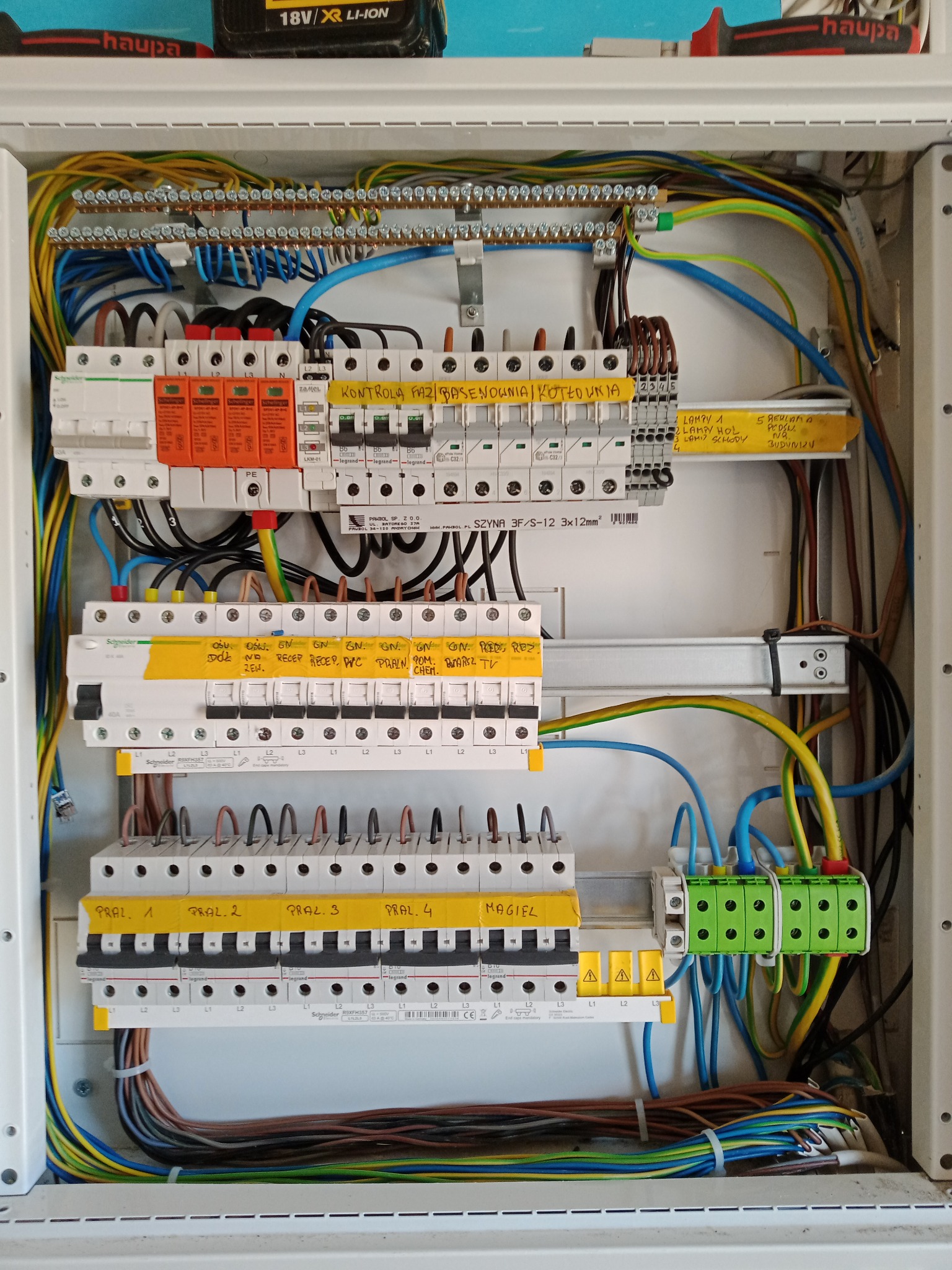 Otwarta skrzynka elektryczna z widocznymi zabezpieczeniami, przewodami i żółtymi etykietami opisującymi obwody, w tym 'Kontrola faz', 'Lampy', 'Pralnia' i 'Magiel'.