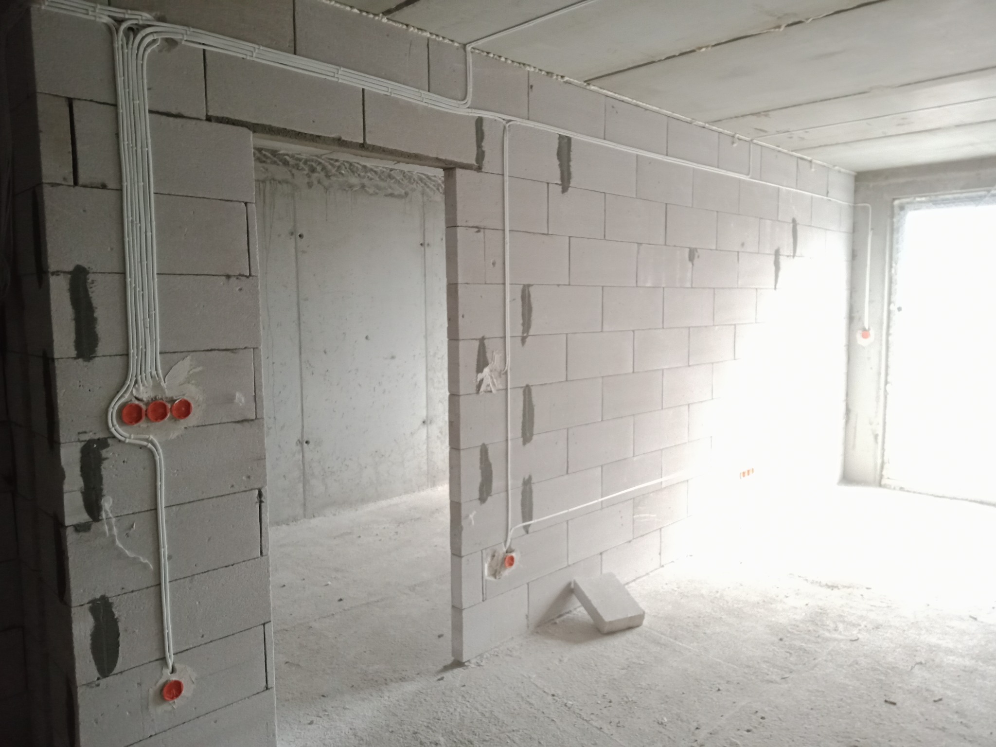 Surowy stan: Białe kable elektryczne prowadzone po ścianach z białych bloczków w nowym, niezamieszkałym jeszcze pomieszczeniu. Widoczne puszki elektryczne w kolorze pomarańczowym. Przewody ułożone...