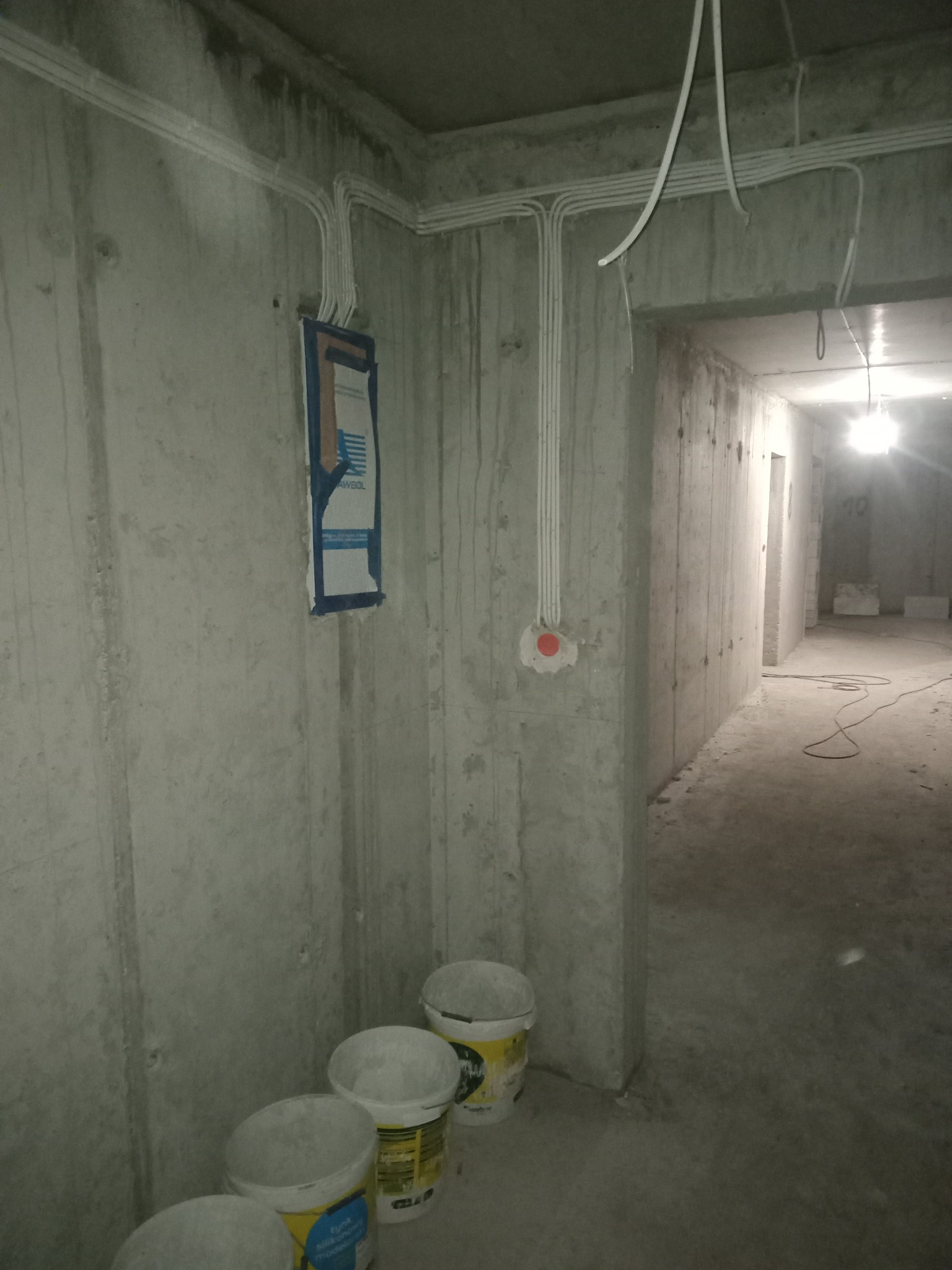 Surowe wnętrze w trakcie prac elektrycznych: widoczne kable prowadzone po ścianach, puszka elektryczna z czerwoną osłoną, skrzynka bezpiecznikowa zabezpieczona taśmą, oraz wiaderka z farbą...