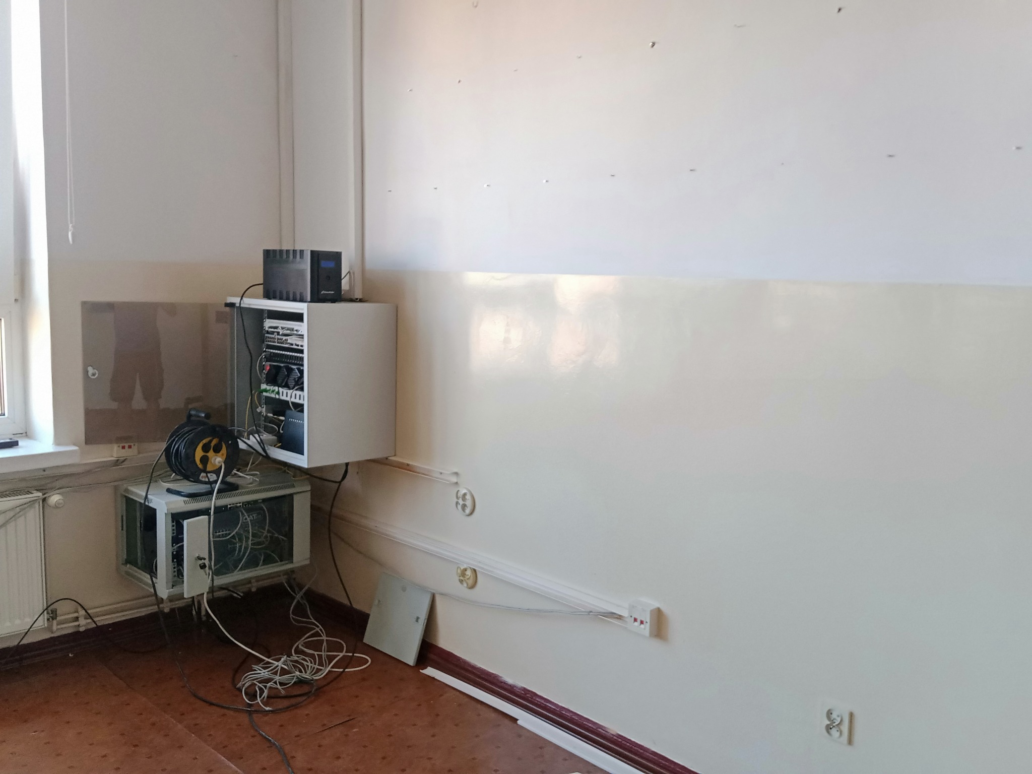 Rozłożona szafa rack z okablowaniem, widoczny UPS i zwinięty przedłużacz na tle ściany z instalacją elektryczną w trakcie montażu.