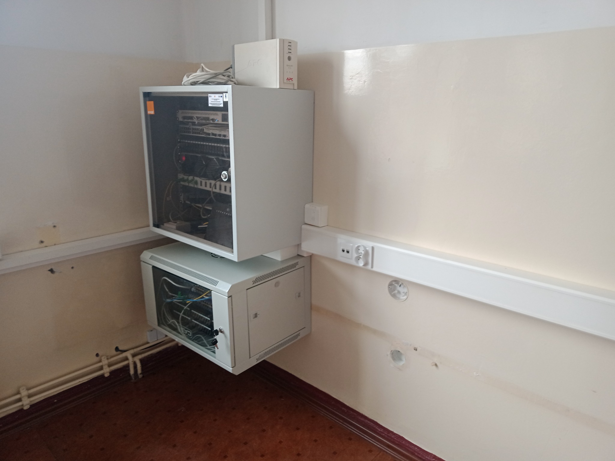 Dwie szafy rack zamontowane na ścianie w pomieszczeniu, z widocznymi kablami i urządzeniami wewnątrz, obok listwy z gniazdkami elektrycznymi i białego kanału kablowego.