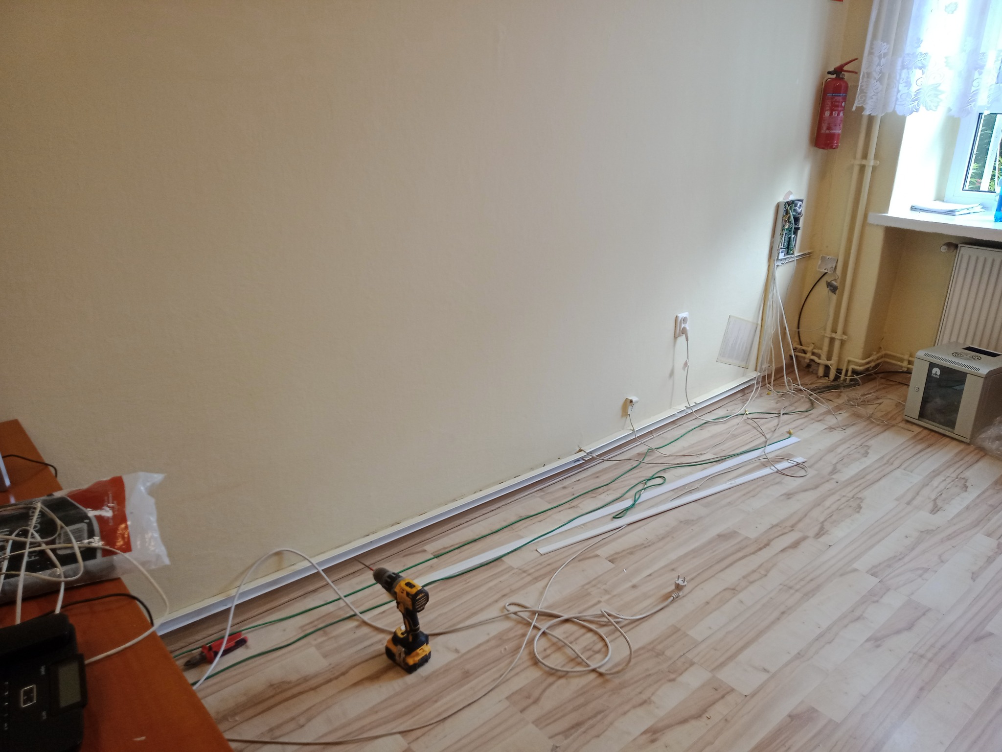 Rozłożone kable elektryczne na podłodze podczas prac instalacyjnych, widoczna skrzynka z przewodami w ścianie, wiertarka akumulatorowa leżąca obok, listwy przypodłogowe i gniazdka elektryczne w tle.