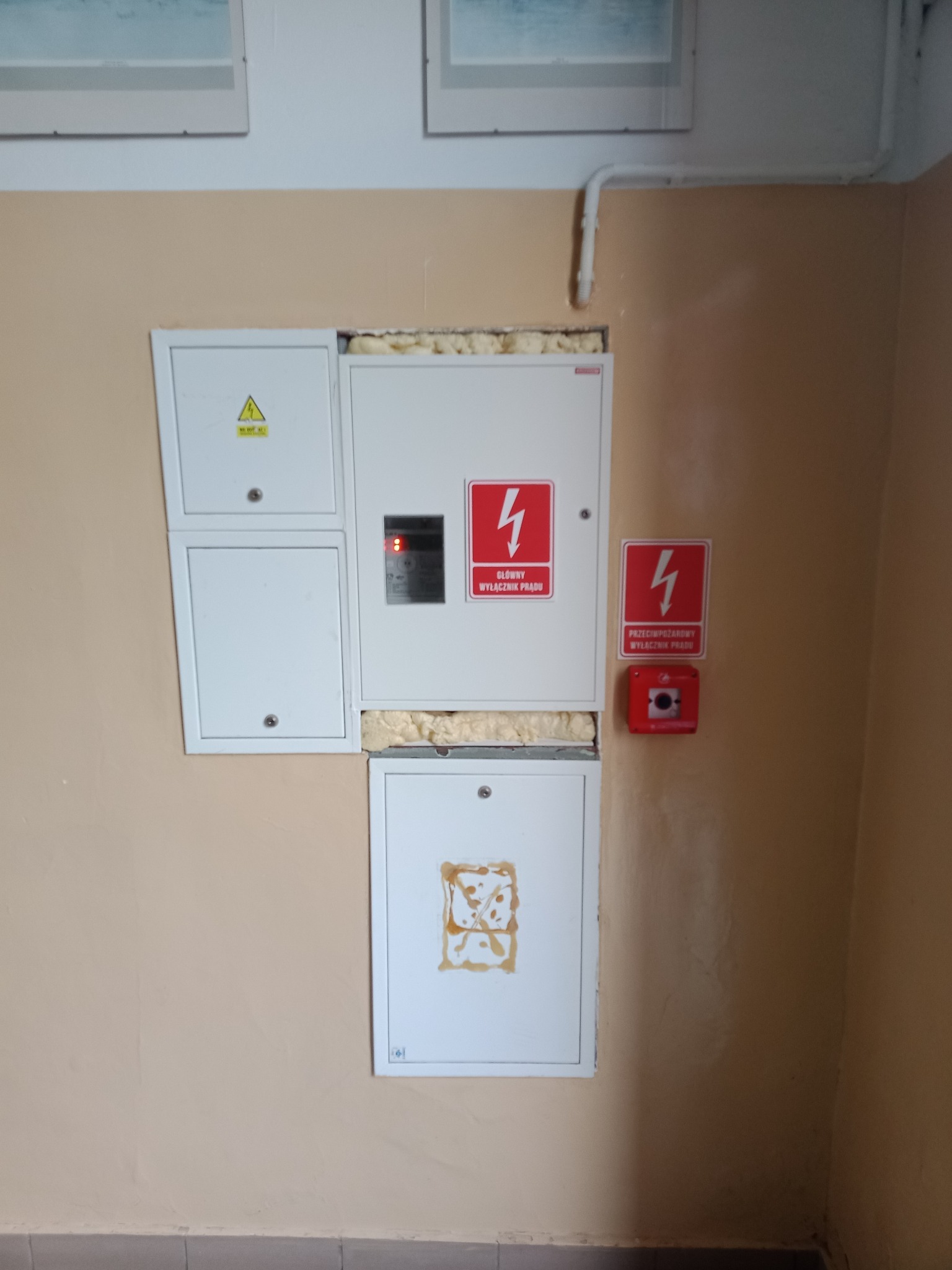 Tablica rozdzielcza z widocznymi zabezpieczeniami, głównym wyłącznikiem prądu i przyciskiem przeciwpożarowym, zamontowana na beżowej ścianie wewnątrz budynku.
