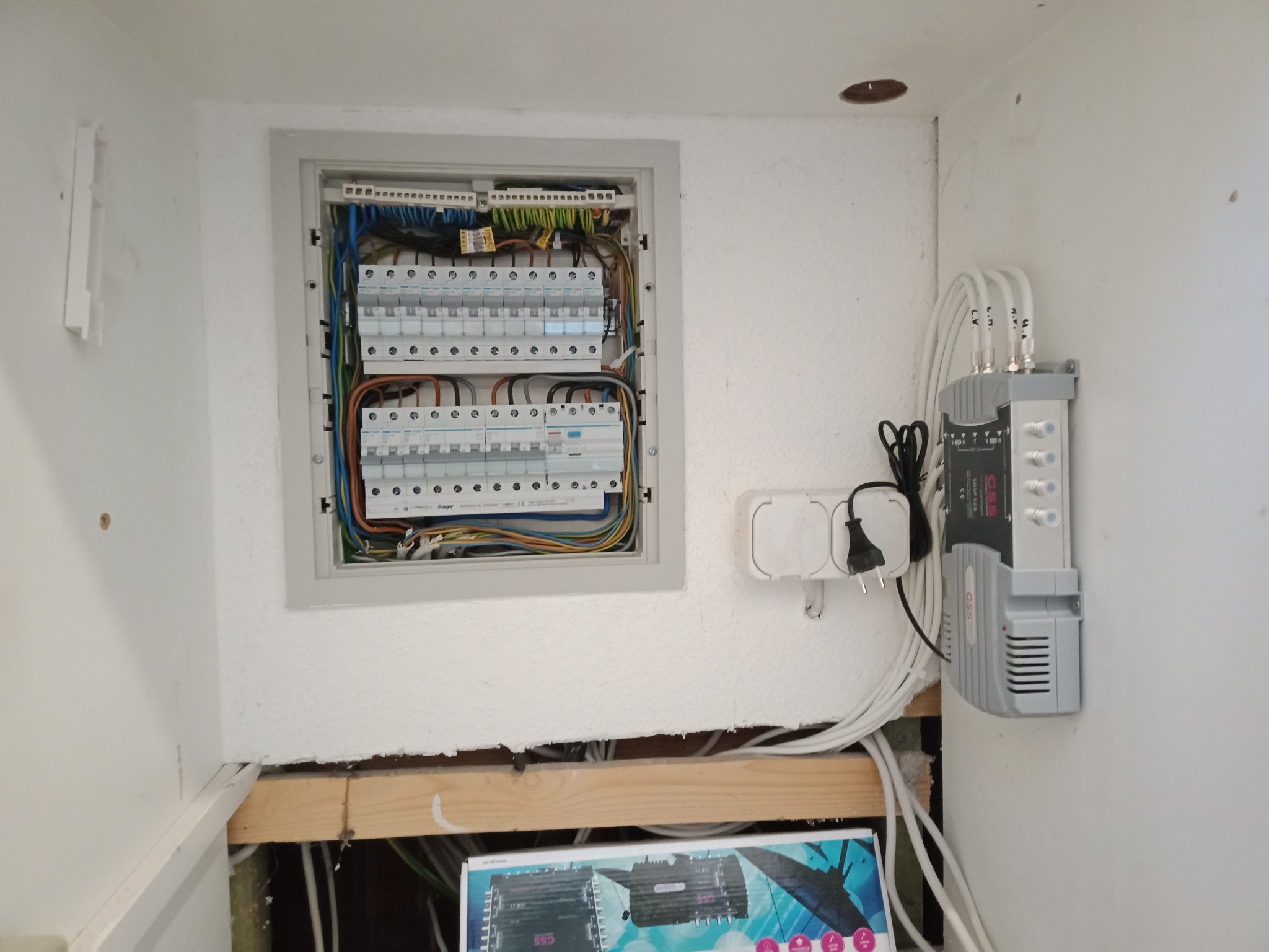 Otwarta skrzynka elektryczna z bezpiecznikami i okablowaniem na białej ścianie, obok gniazdko elektryczne i wzmacniacz sygnału z kablami koncentrycznymi.