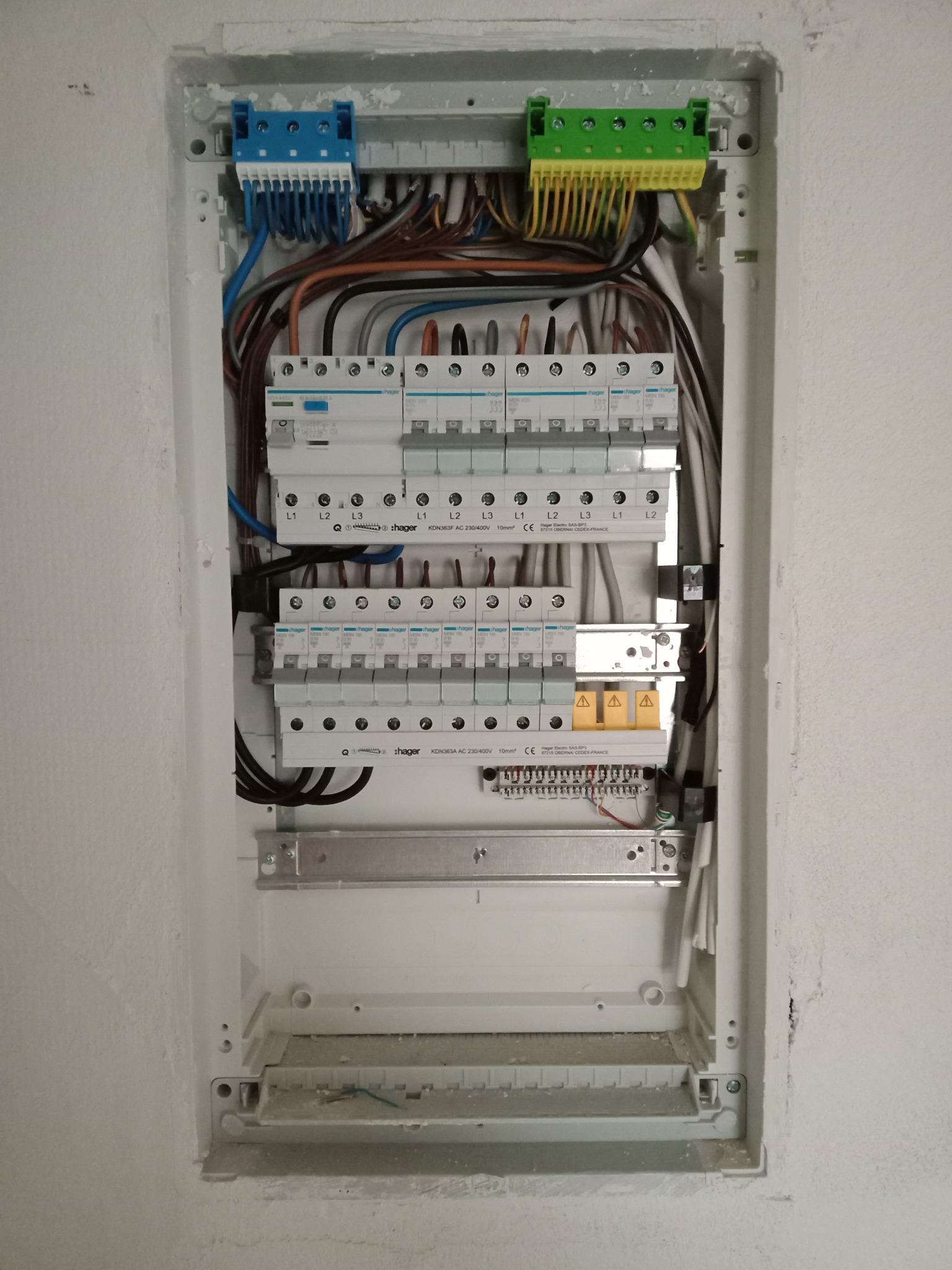 Otwarta skrzynka elektryczna z zainstalowanymi bezpiecznikami, przewodami i złączkami, widoczne oznaczenia L1, L2, L3 oraz logo Hager na bezpiecznikach.