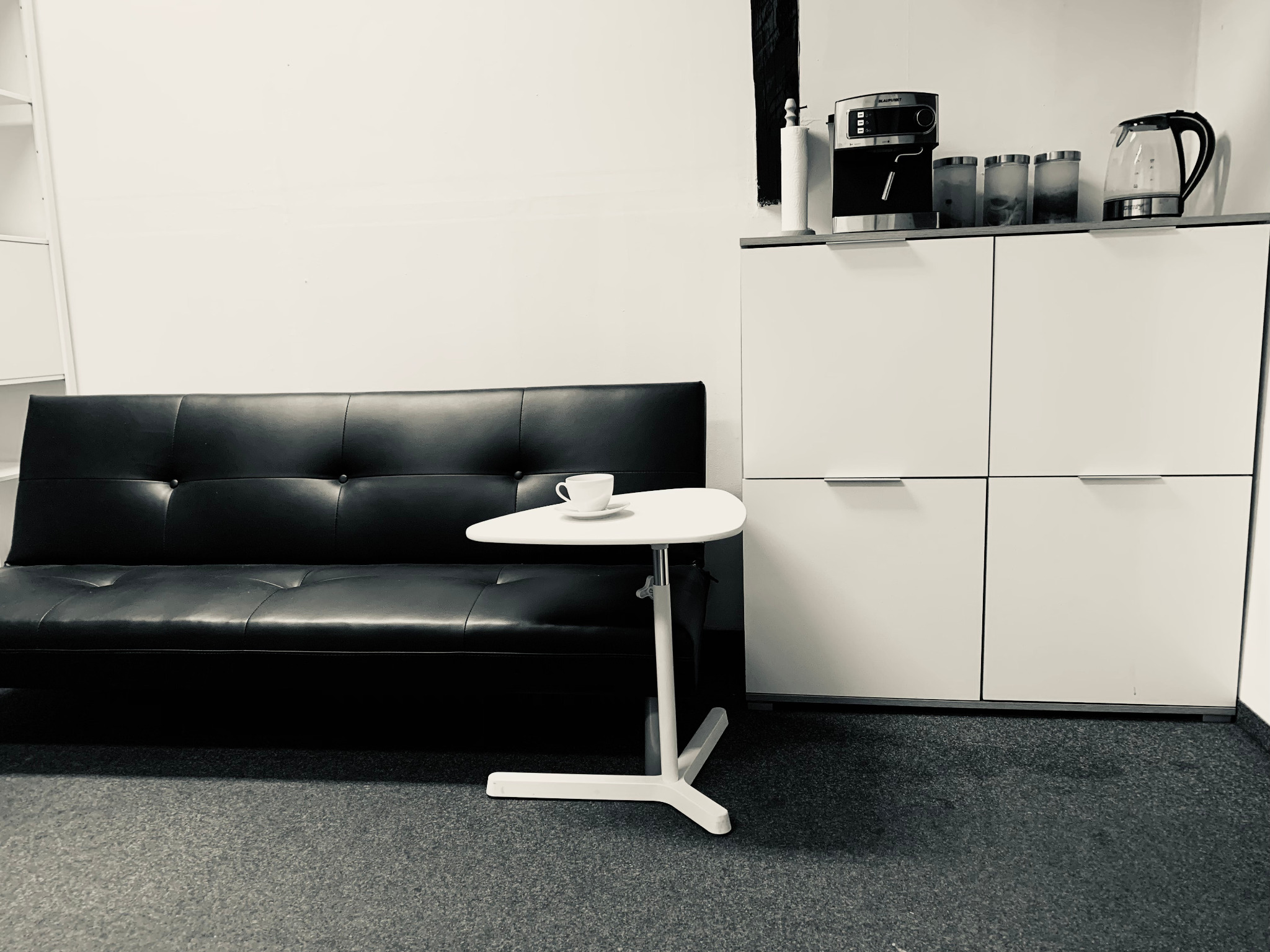 Minimalistyczne biuro z czarną sofą, białym stolikiem z filiżanką i szafką z ekspresem do kawy. Stonowana kolorystyka, proste linie, nowoczesny design.