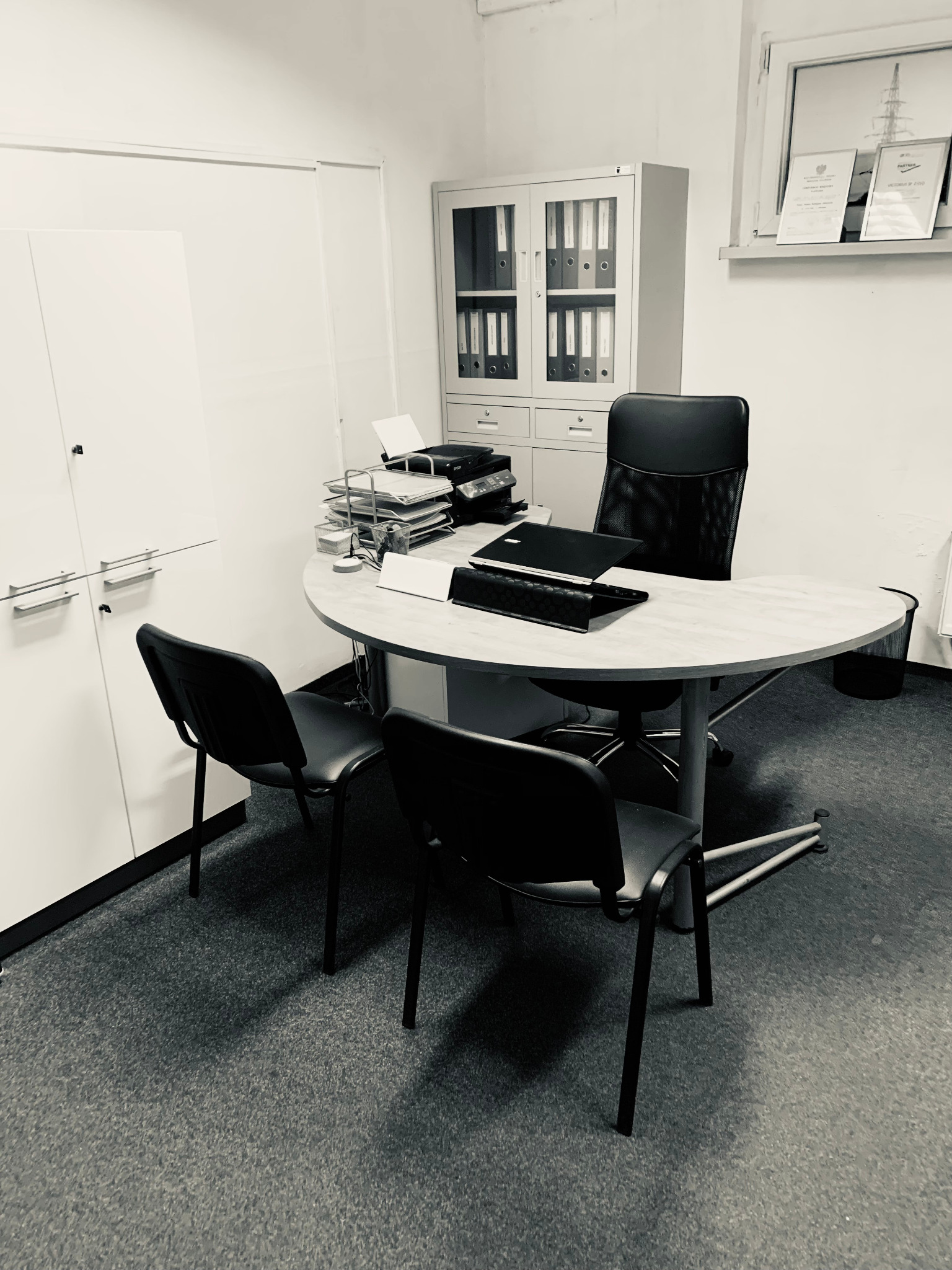 Biuro z okrągłym stołem, krzesłami i szafą na dokumenty. Na ścianie dyplomy. Stonowana kolorystyka. Widok na uporządkowane miejsce pracy.